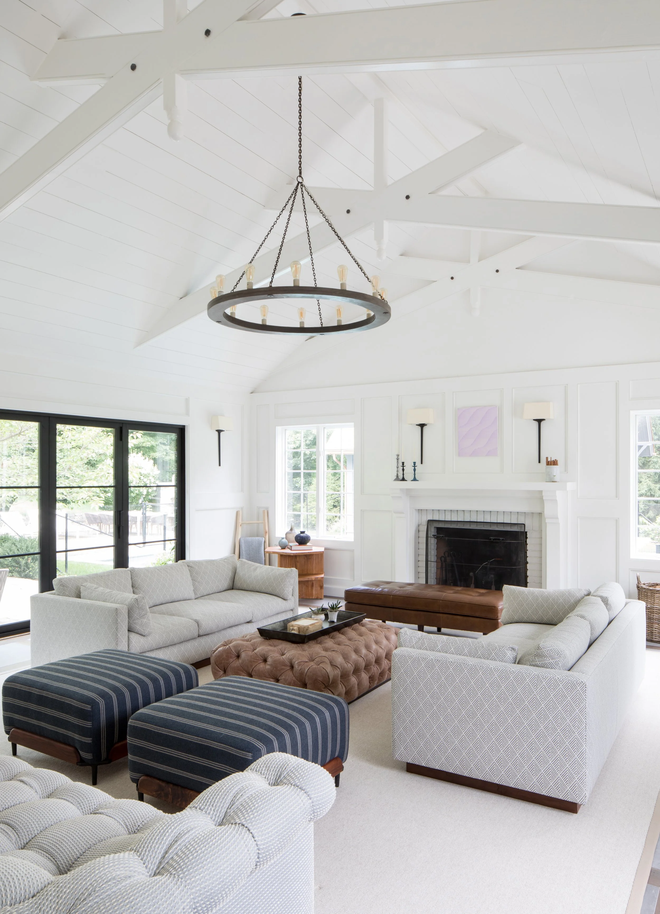 Living room with white walls and ceiling, black chandelier, beige and blue furniture, white couches, brown tufted ottoman, and large windows.
