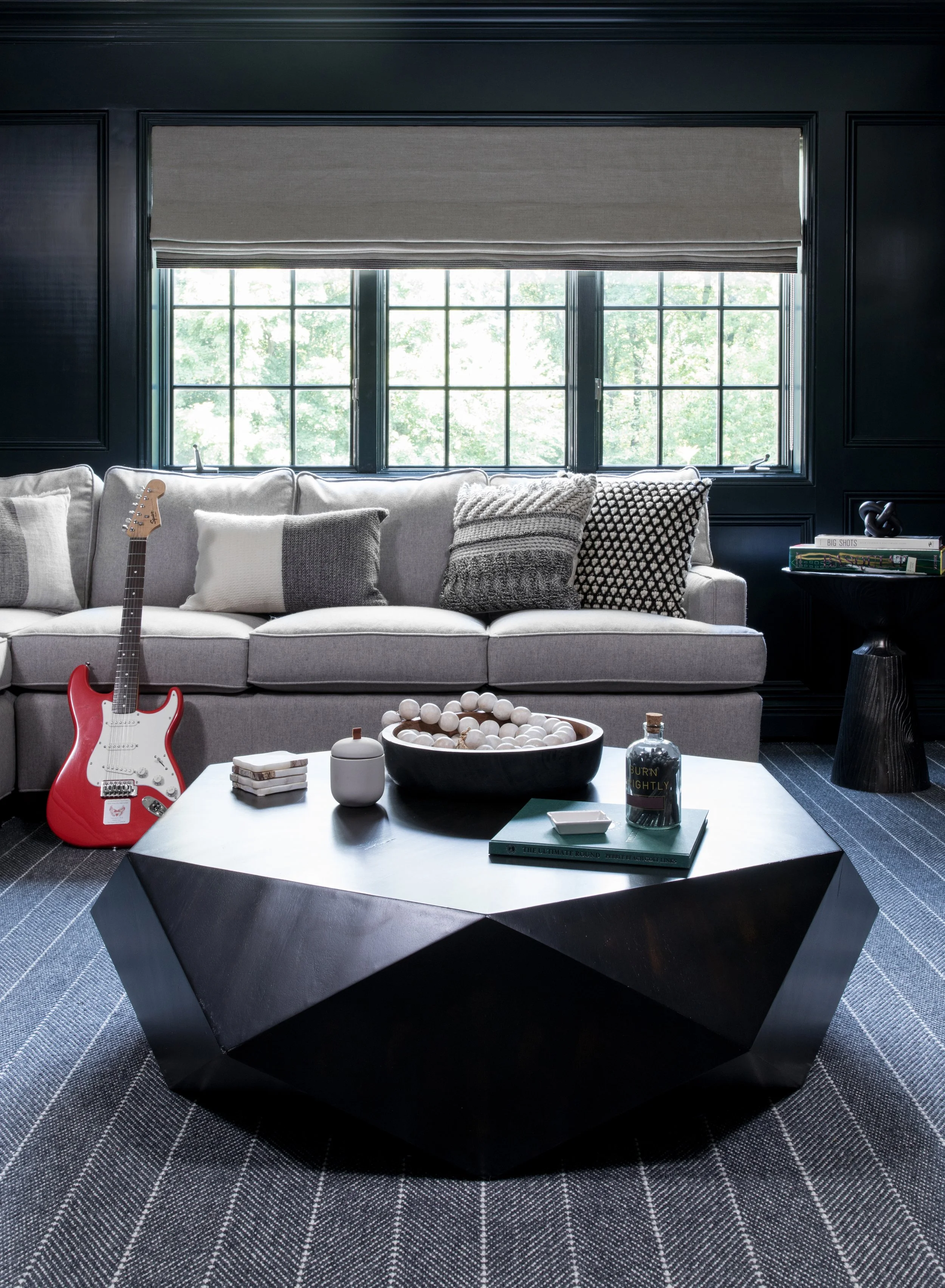 Modern living room with a light gray sofa, patterned pillows, a black geometric coffee table, a red electric guitar, and a window with black framing letting in natural light.