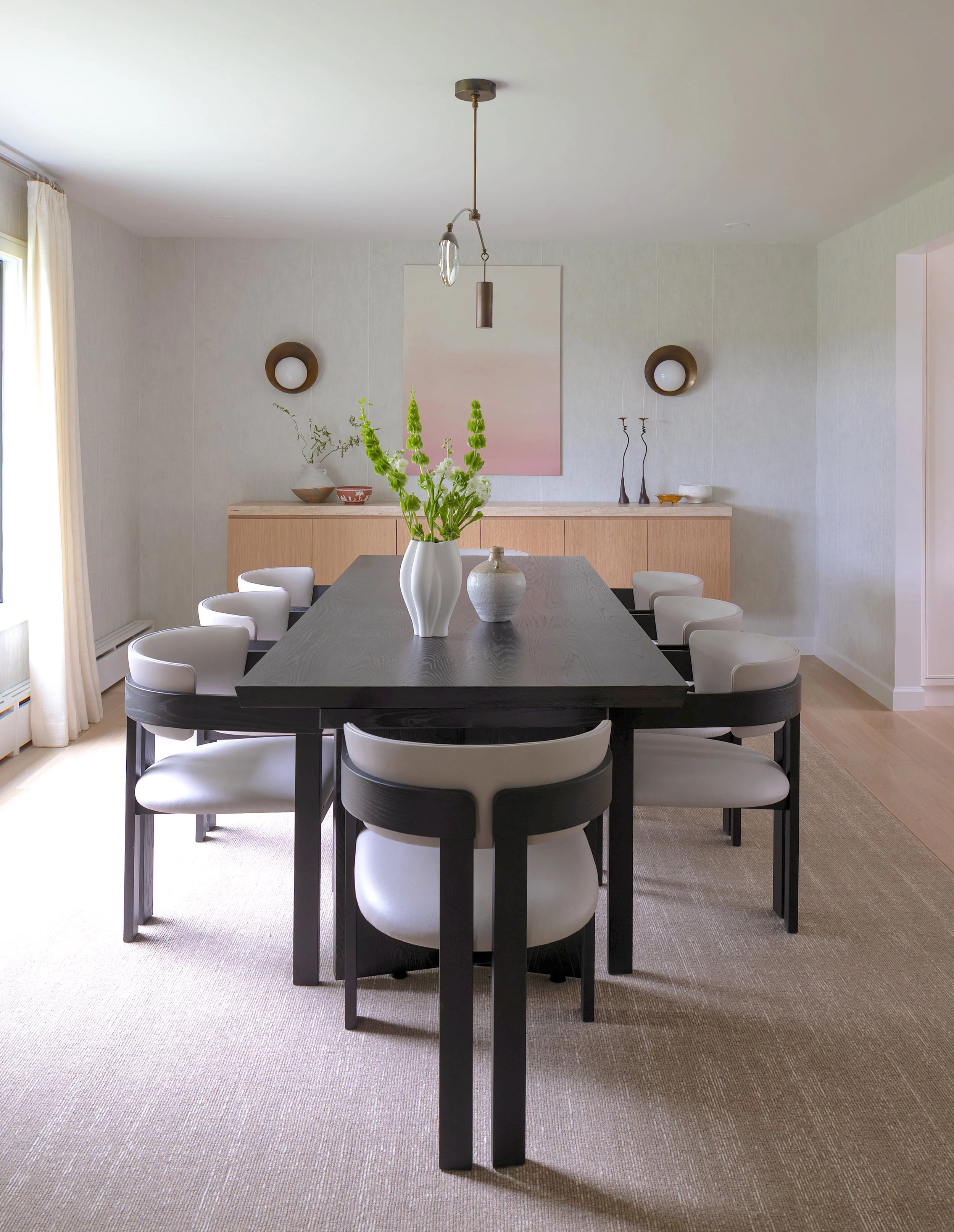 Modern dining room with a black wood table, white upholstered chairs, a beige rug, a sideboard with decorative objects, a large pink and white abstract painting, and green floral centerpiece in a white vase.