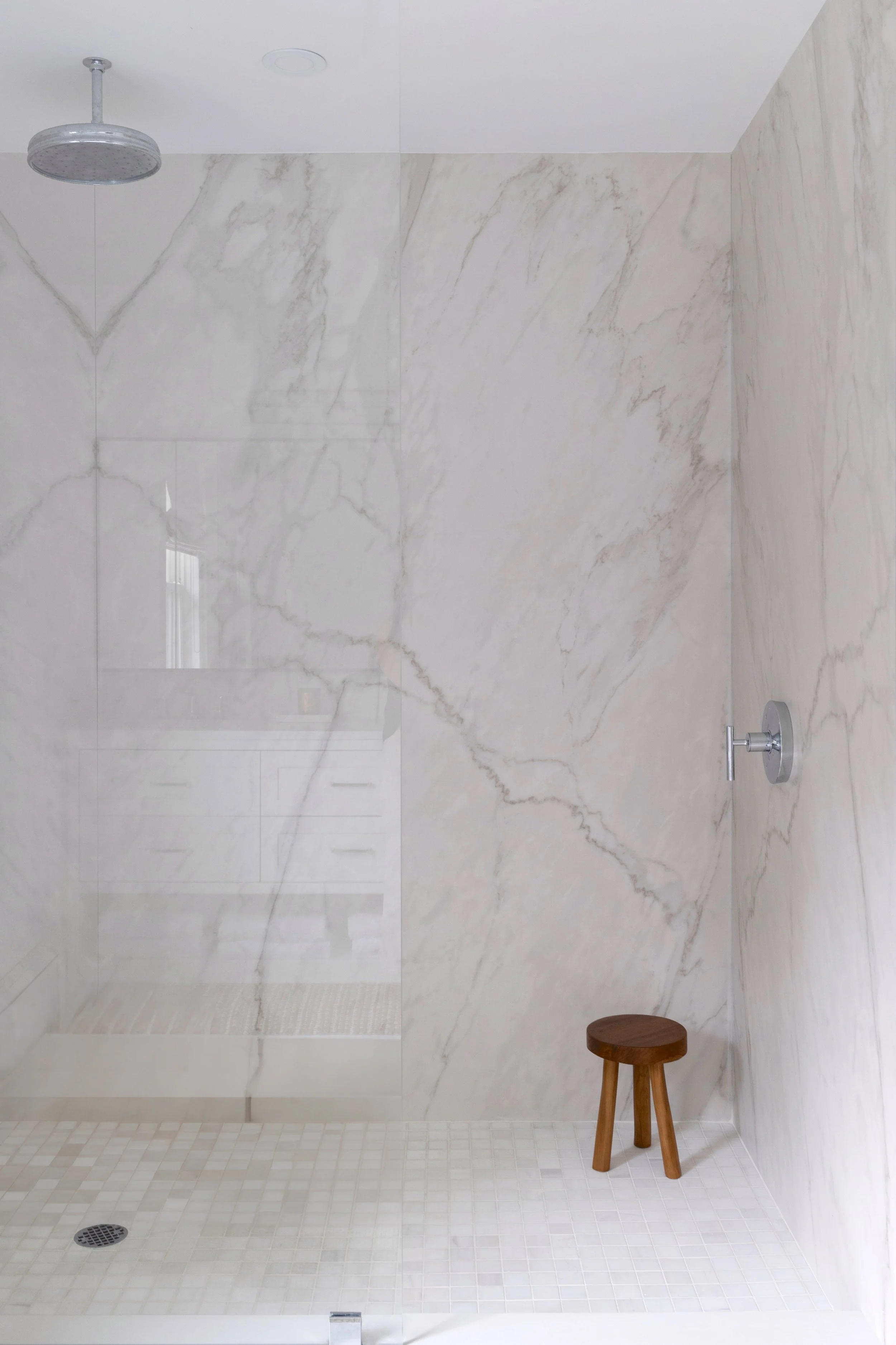 Minimalist walk-in shower with large marble tiles, a round showerhead, a small wooden stool, and a glass shower enclosure.