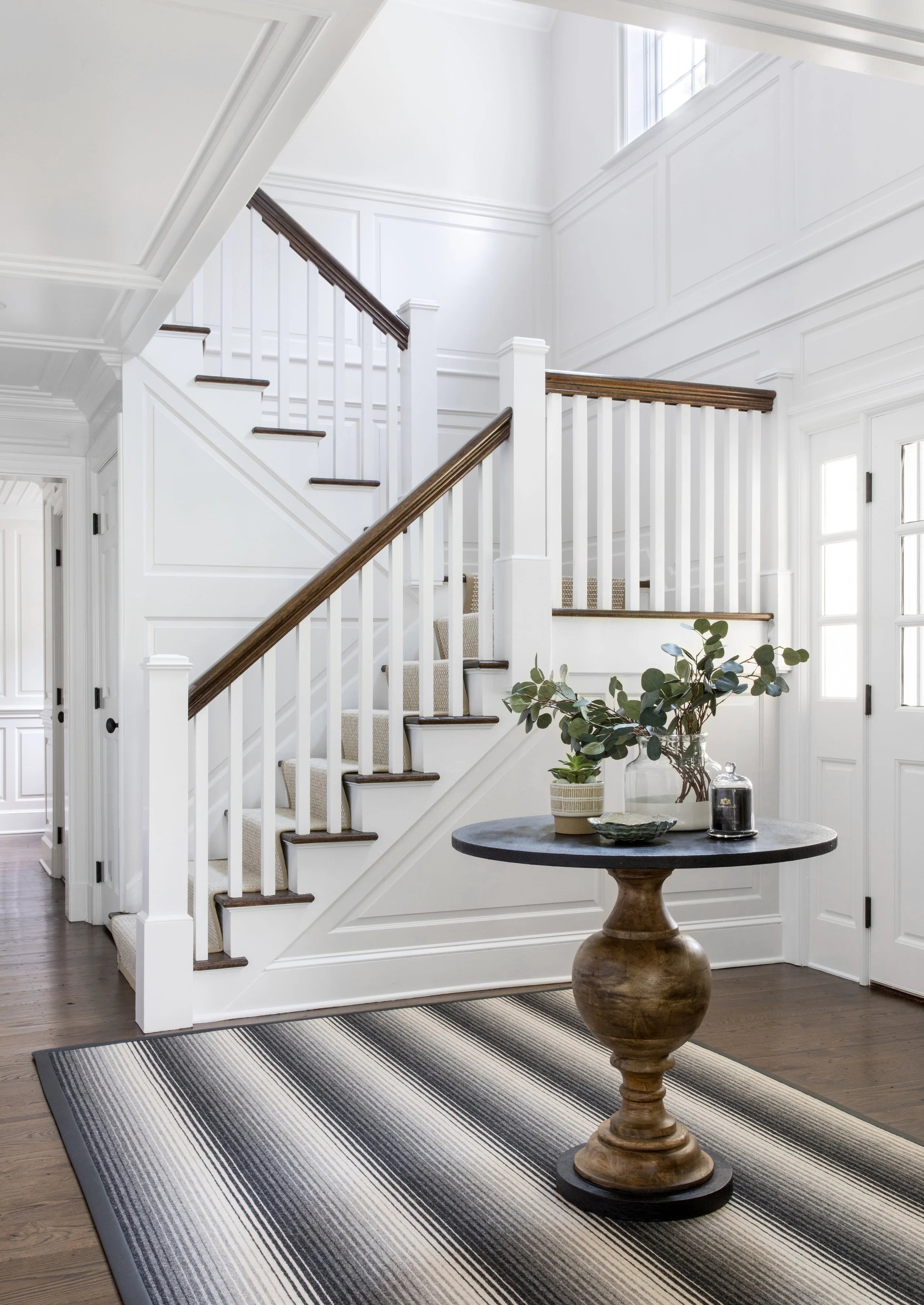 White entryway with staircase, wooden flooring, black and white striped rug, round wooden table with green potted plant, lamp, vase, and decorative objects.