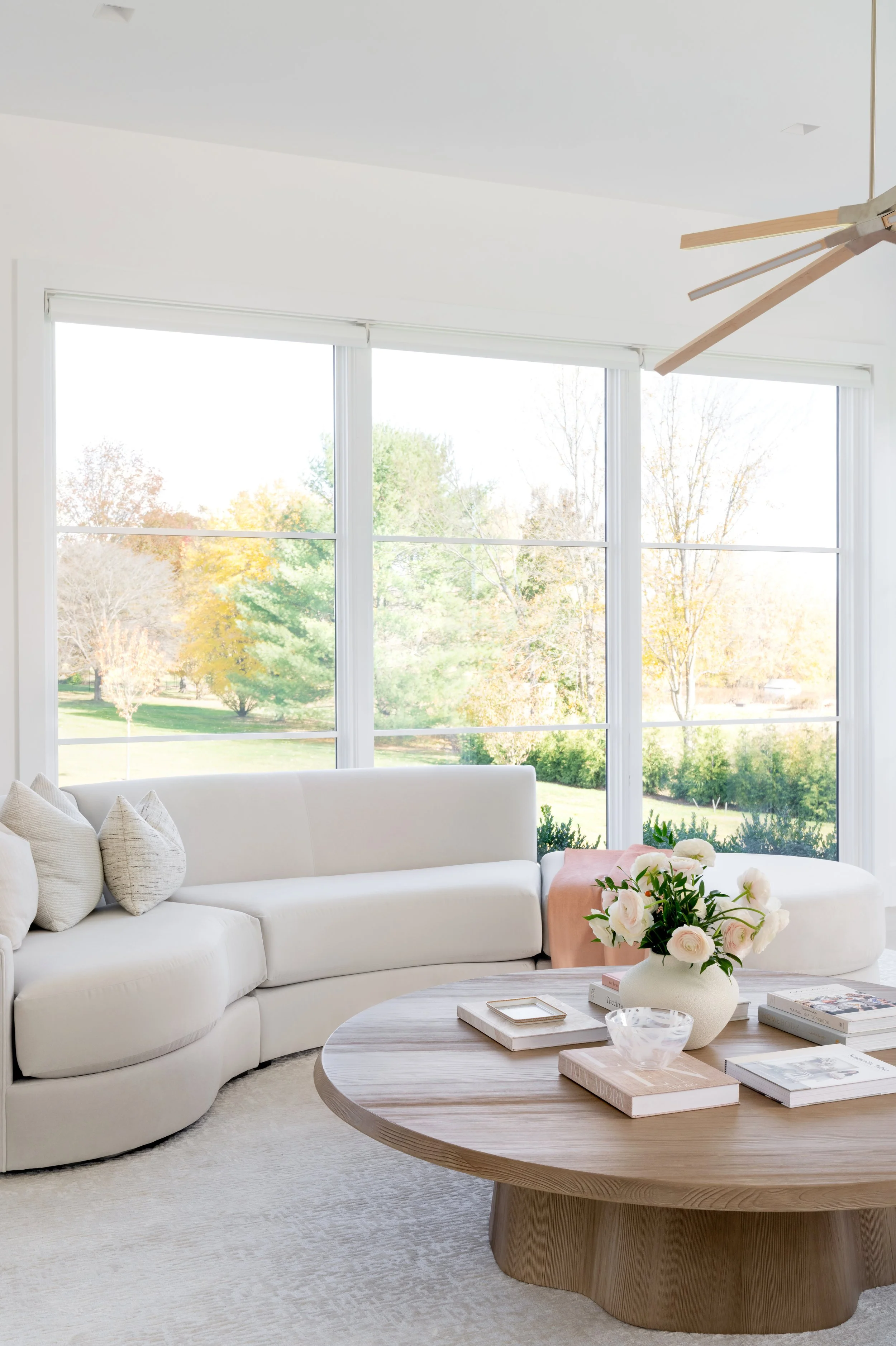 Bright living room with large windows, white sectional sofa, round wooden coffee table with books, a vase of white flowers, and a modern ceiling fan.