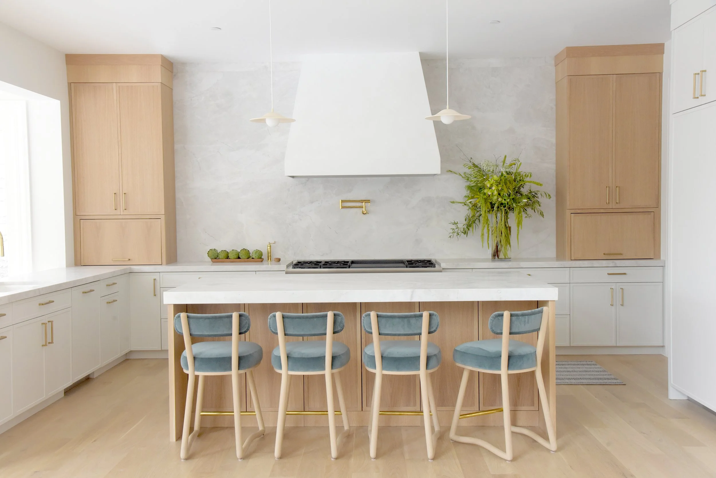 Modern kitchen with white and light wood cabinetry, marble countertops, and bar stools with blue cushions at an island. Green plants and brushed gold accents are visible.