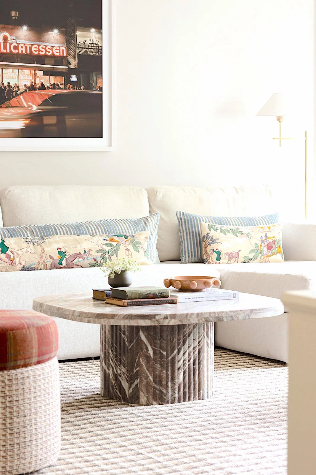 Living room with a white sofa decorated with patterned pillows, a round marble coffee table with books, a small flower vase, and a decorative bowl, a multicolored upholstered ottoman, a striped rug, and wall art depicting a city scene with a sign tha