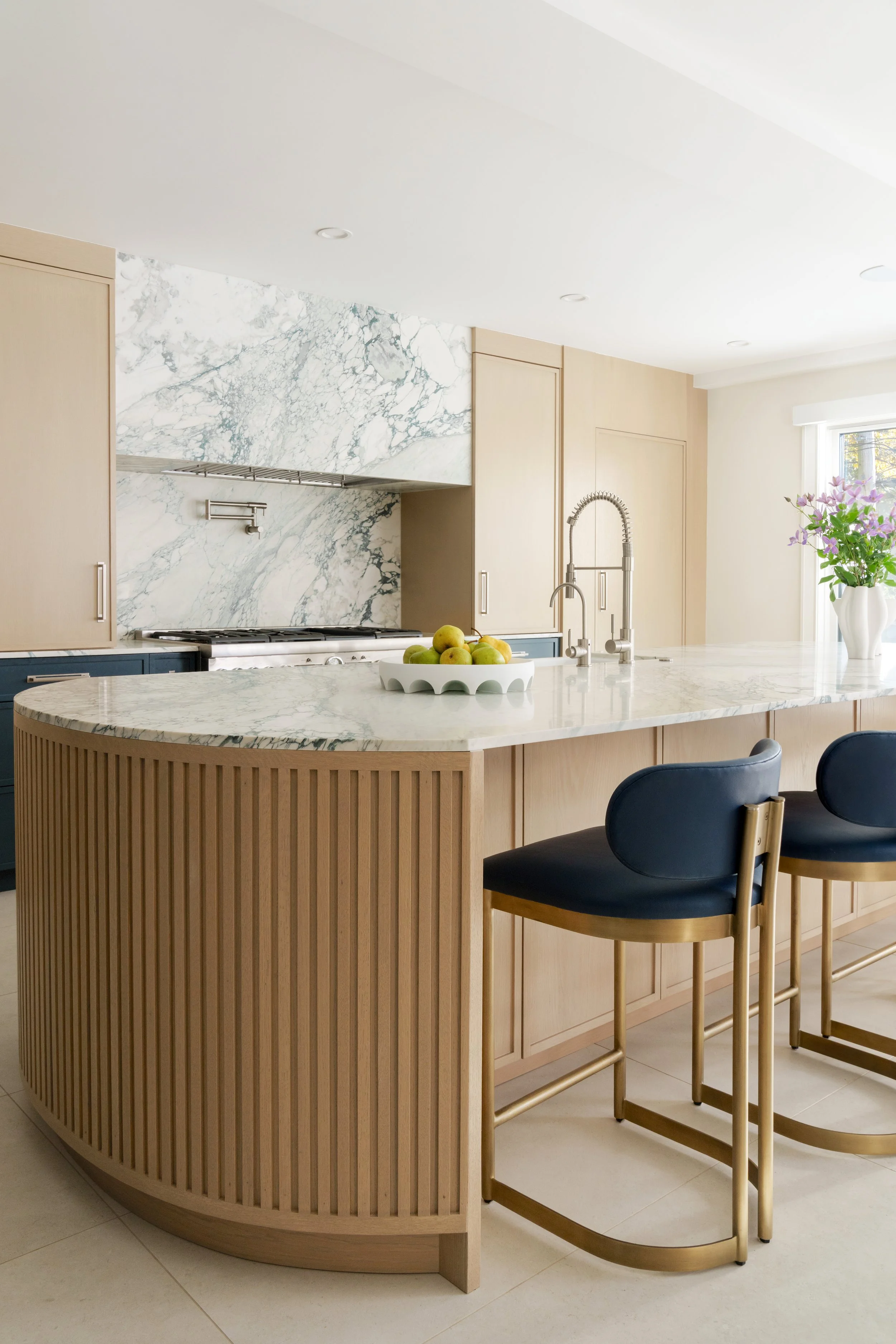 Modern kitchen with a marble island, light wood cabinetry, and black barstools, featuring a vase with purple flowers near a window.
