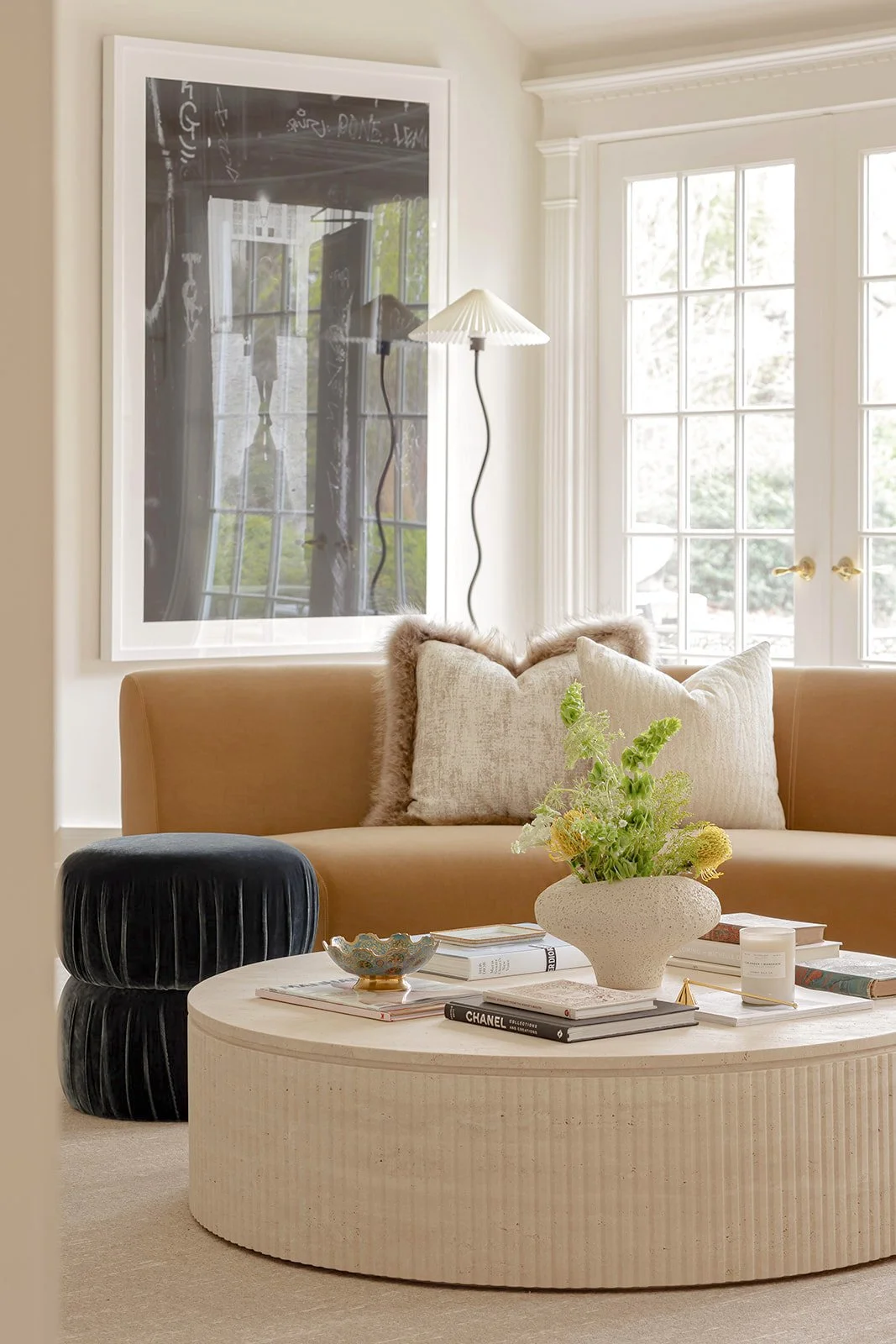 Living room with beige sofa, black velvet ottoman, and round coffee table with books, a vase of green plants, and a decorative bowl. Large windows with white trim and a floor lamp with a white pleated shade.