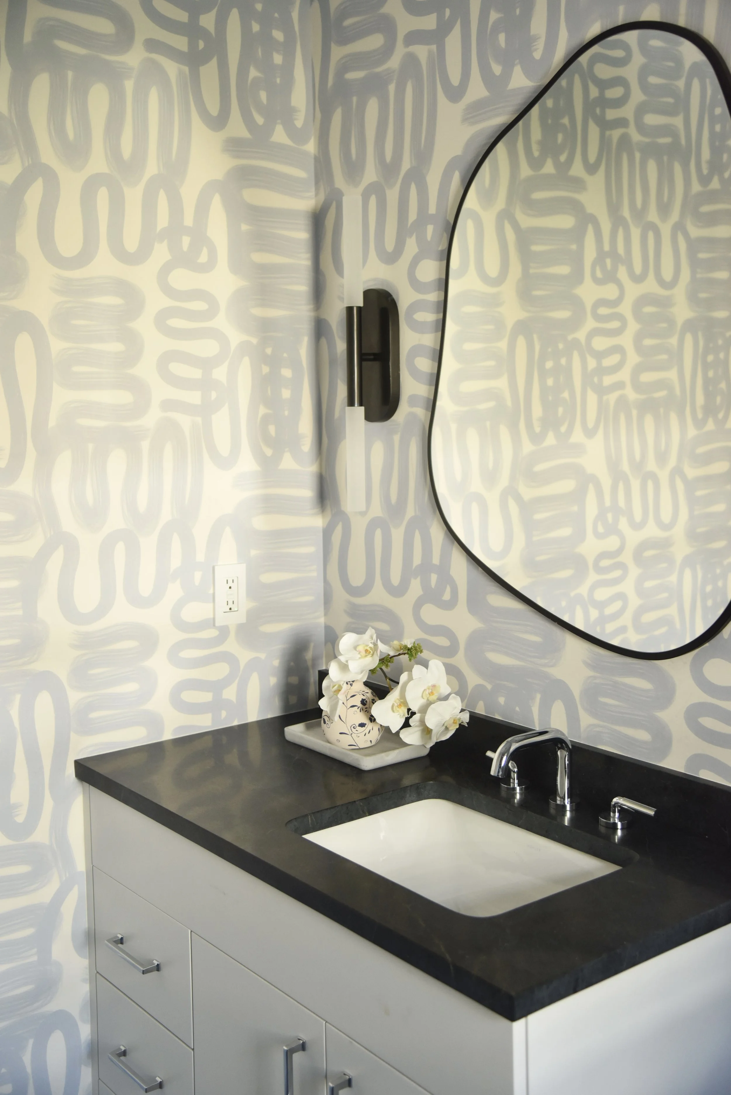 A bathroom vanity with a white cabinet and black countertop, a rectangular sink, and a silver faucet. A decorative ceramic vase with white flowers sits on the counter, and a large, rounded mirror hangs above. The wall has a patterned wallpaper with g