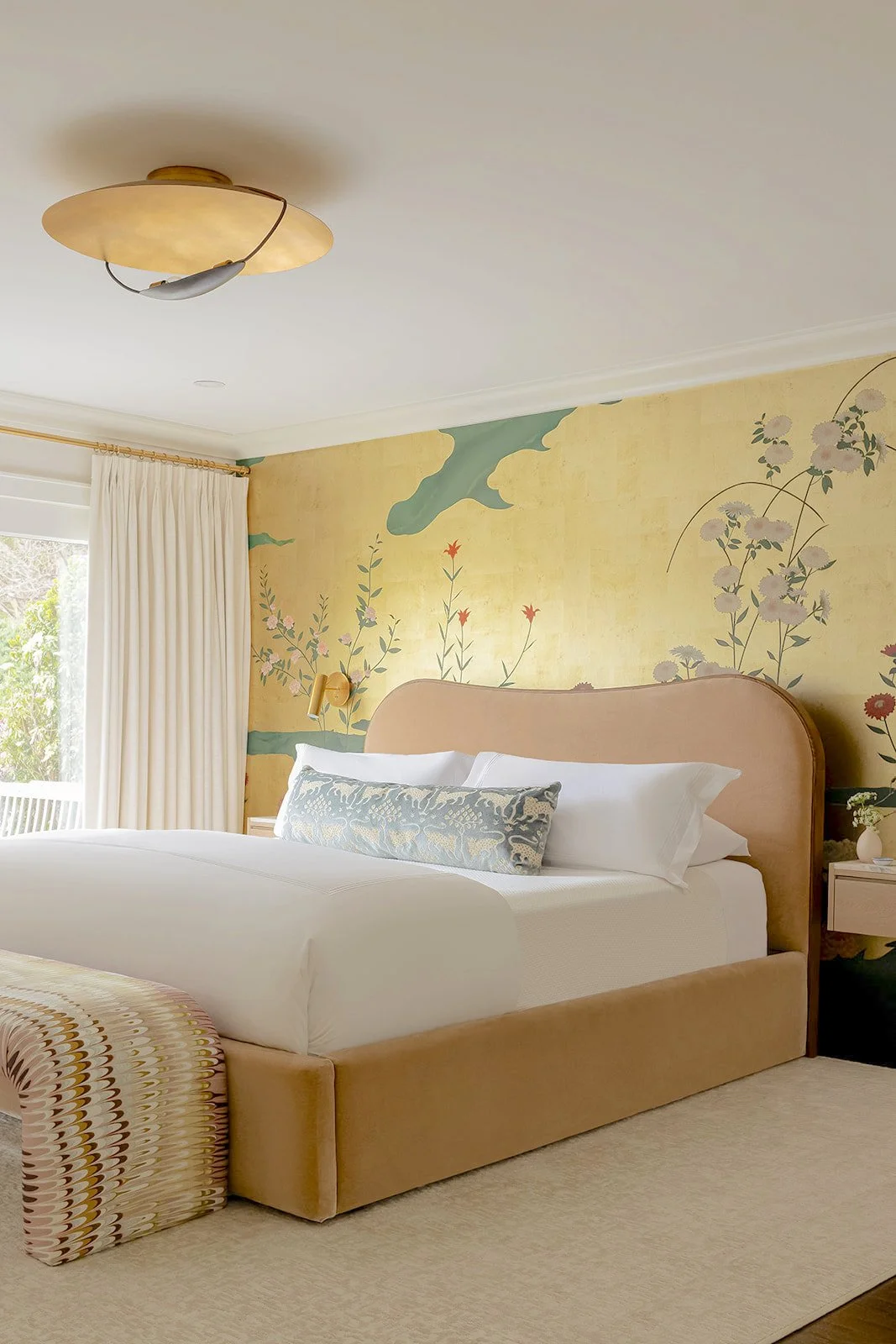 A bedroom with a bed, pillows, a mural wall with floral and cloud designs, a window with cream curtains, and a ceiling light fixture.