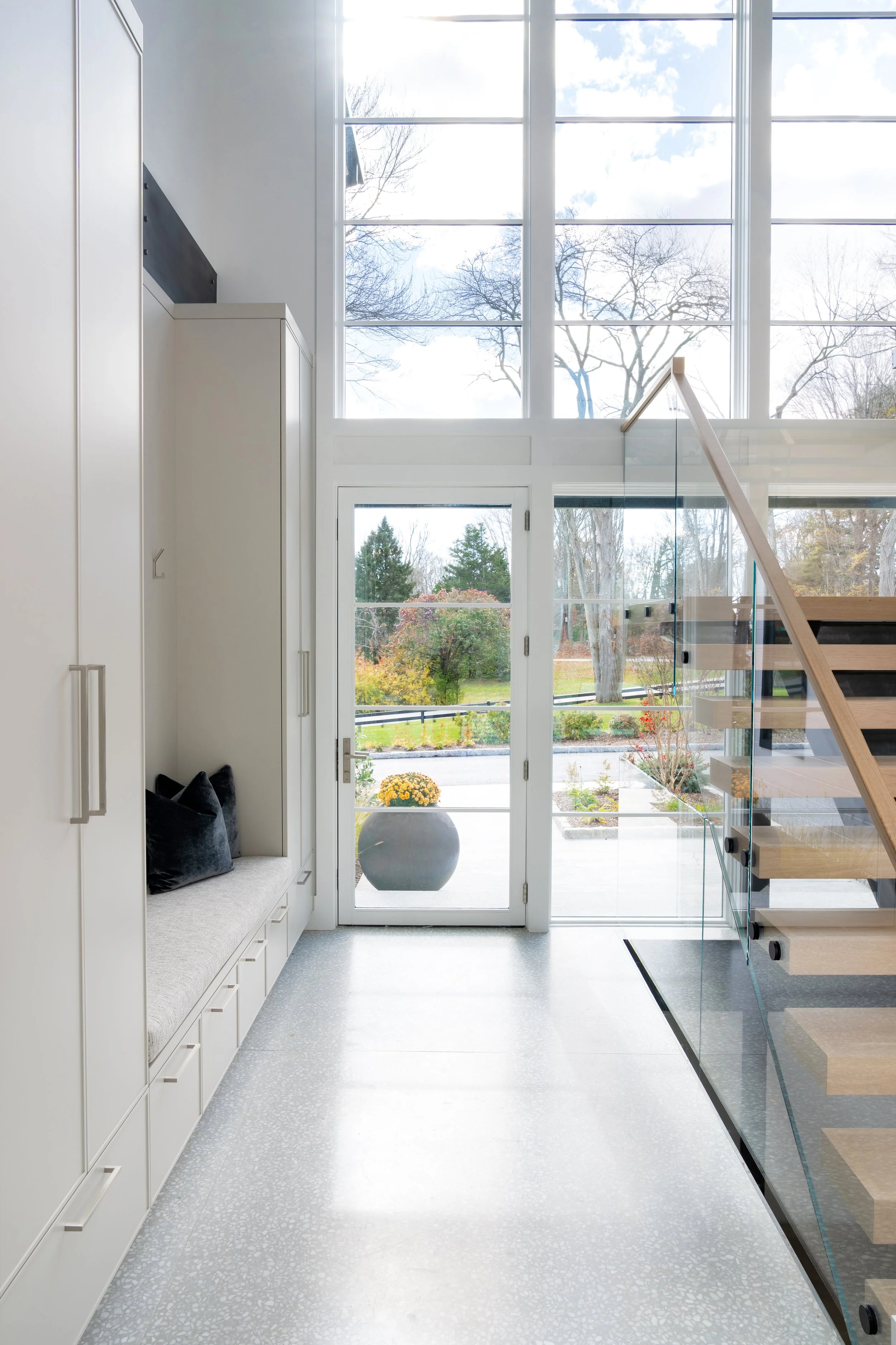 A modern minimalist interior with a staircase, glass railing, white cabinetry with drawers, black pillows on a bench, large windows letting in natural light, and a view of a landscaped outdoor area with trees and flowers.