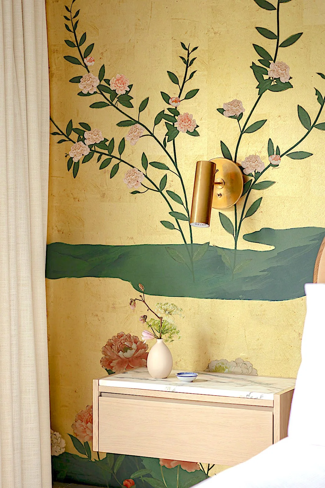 Interior wall with floral mural, gold wall sconce, white dresser with marble top, and small vase with flowers.