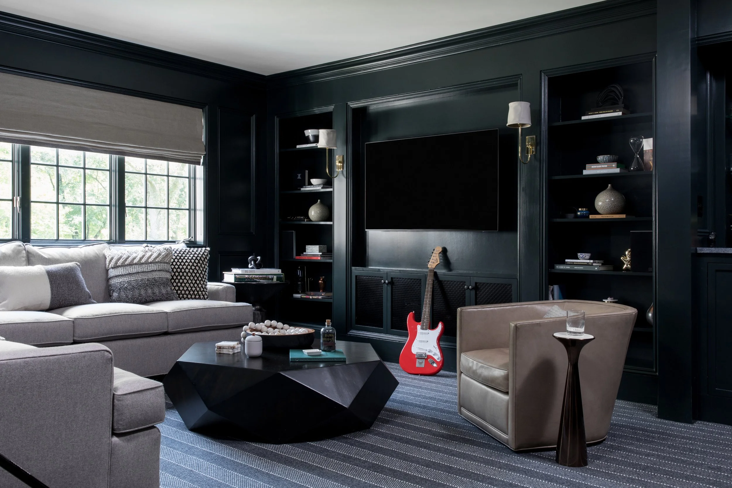 Living room with black walls, large window with beige shades, gray sofa with patterned pillows, black coffee table with decor items, beige armchair with glass of water, electric guitar near black entertainment center, bookshelves with decorative obje