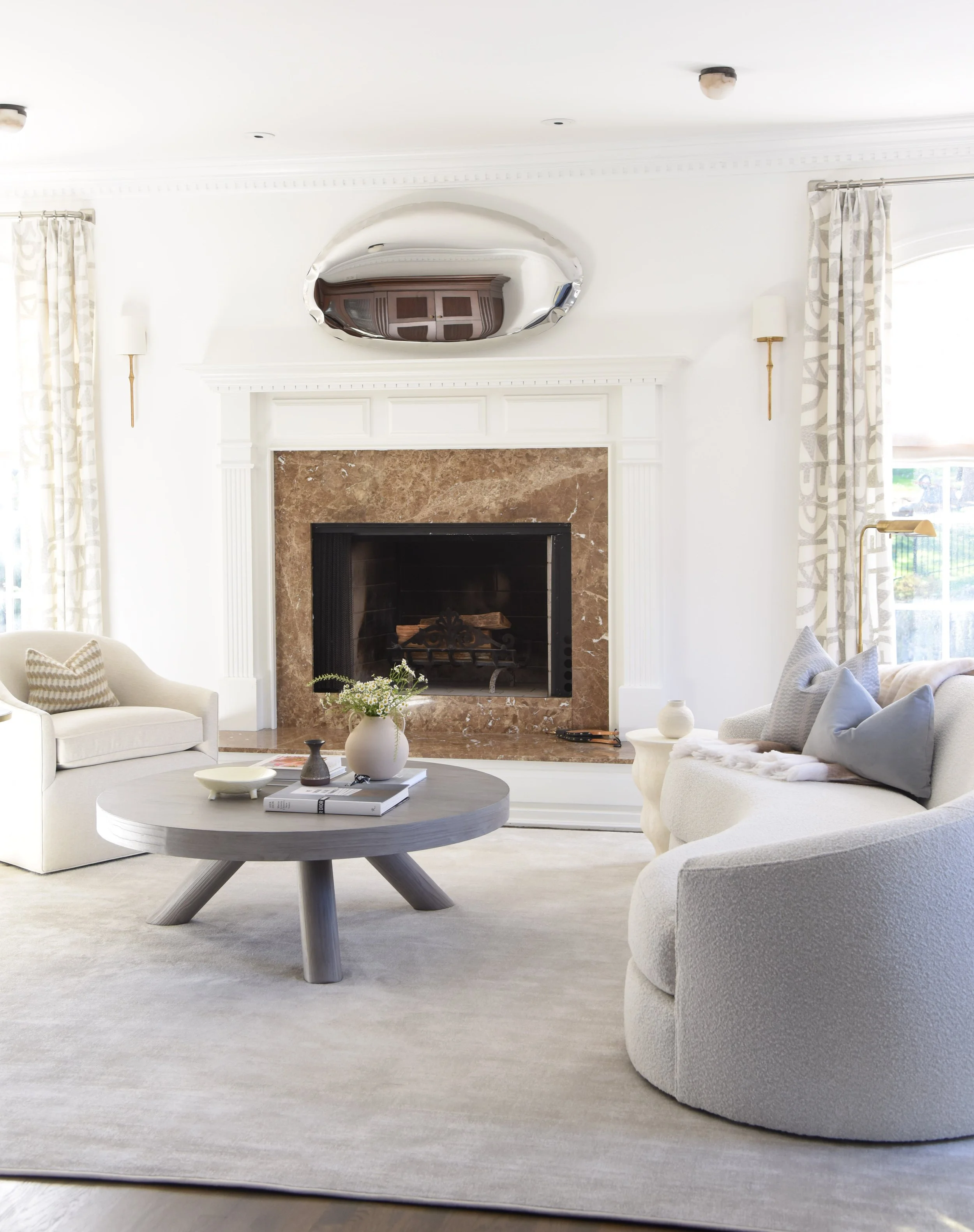 Bright living room with white walls, a fireplace with a brown marble surround, a round wooden coffee table with decorative books and vases, off-white sofas with gray and patterned pillows, large windows with patterned curtains, and a reflective ceili