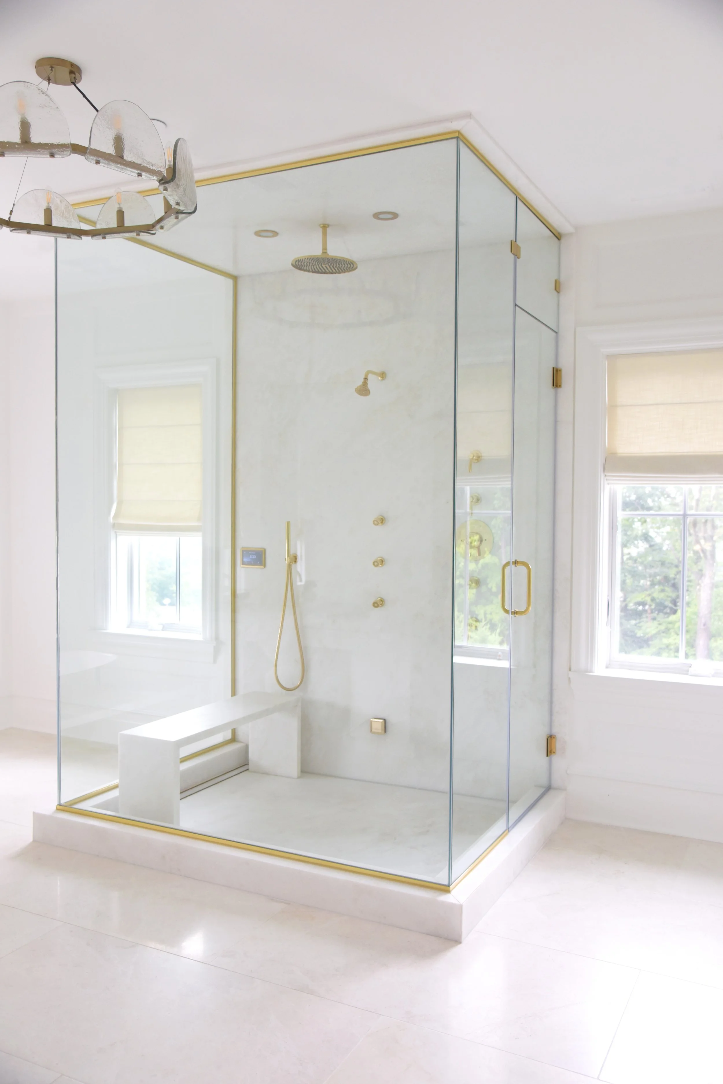 Modern glass shower enclosure with a black handle, gold fixtures, a rain showerhead, and a built-in marble bench in a bright bathroom with windows and white walls.