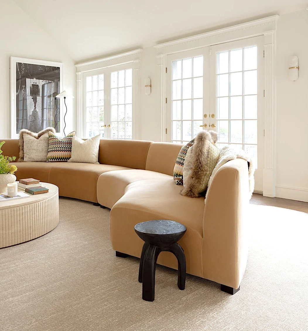 Bright living room with beige sectional sofa, various decorative pillows, round coffee table with books and candles, black side table, large windows, and framed artwork on white walls.
