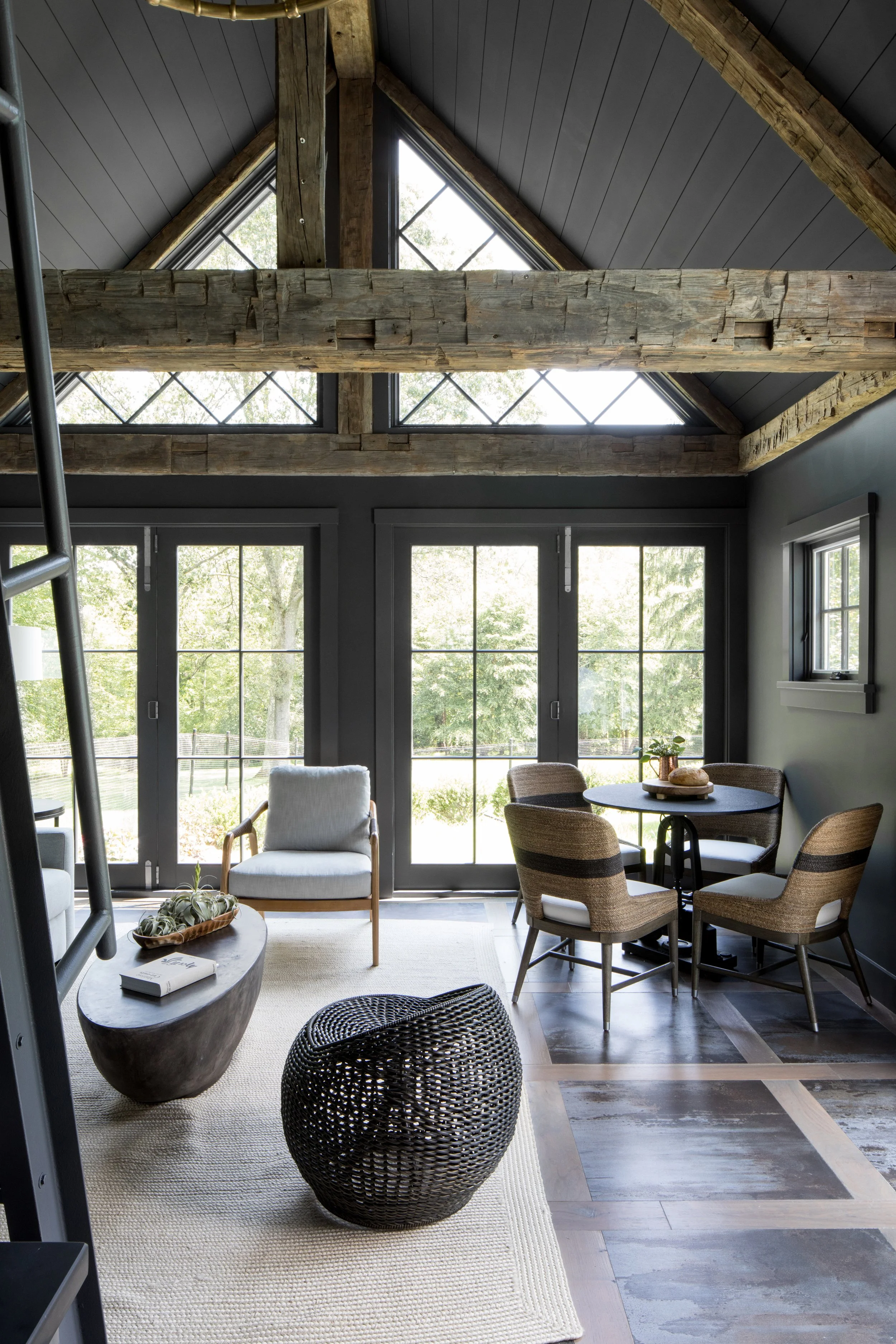 Living room with large glass doors, dark walls, and a vaulted ceiling with wooden beams and triangular windows, furnished with a round table, four woven chairs, an armchair, a black wicker ottoman, a white rug, and decorative plants.