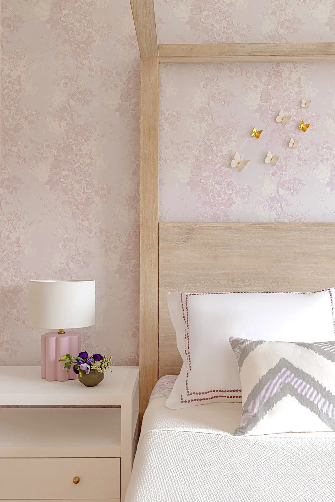 Close-up of a bedroom corner with light wood headboard, white pillow with red stitched border, decorative pillow with chevron pattern, pink and white bedside lamp, small vase with purple flowers, and pink floral textured wall with gold and white butt