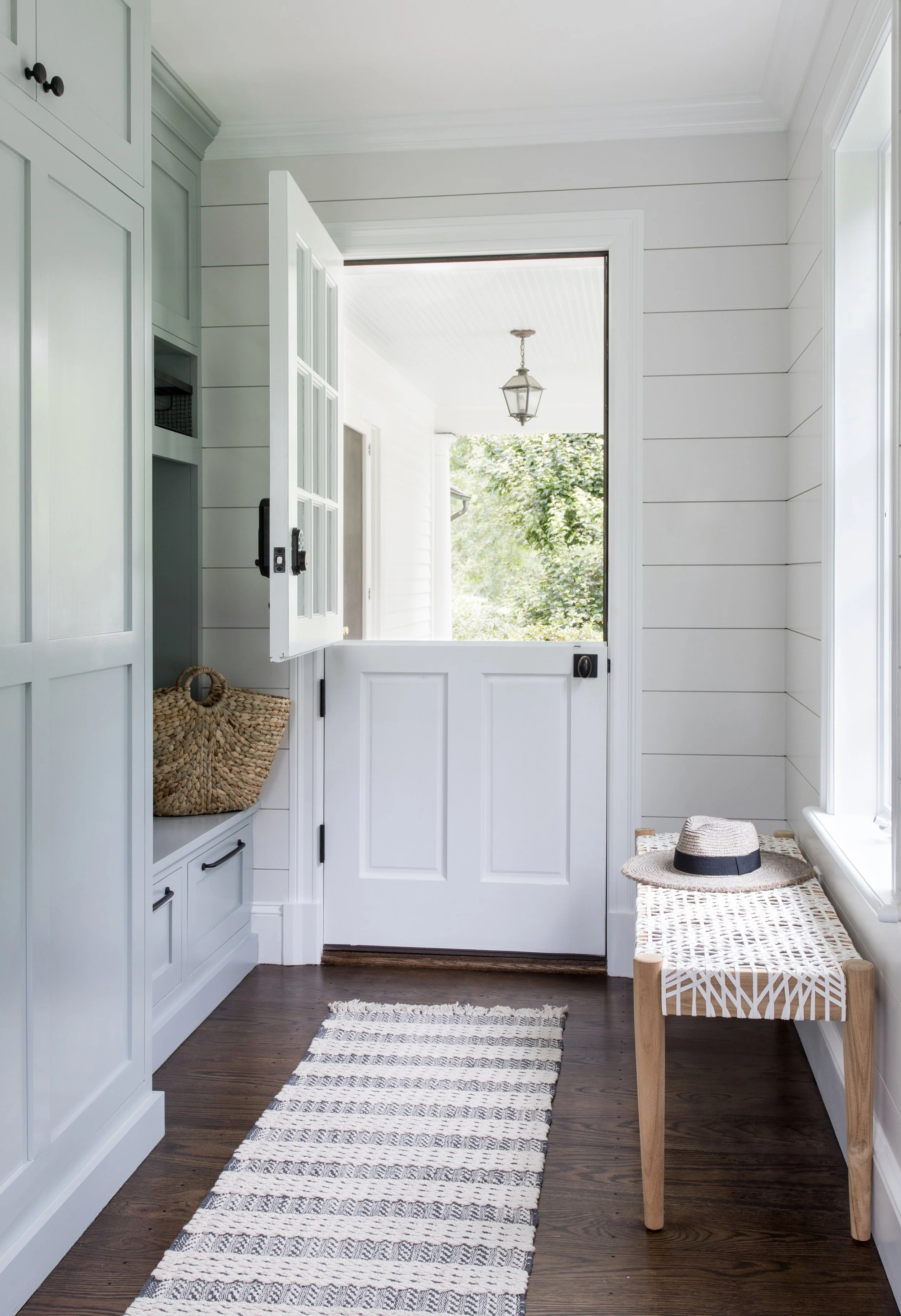 Bright, airy entryway with white shiplap walls, wooden floors, a bench with a hat, a woven basket on a built-in storage unit, and an open door leading outside and revealing a greenery view.