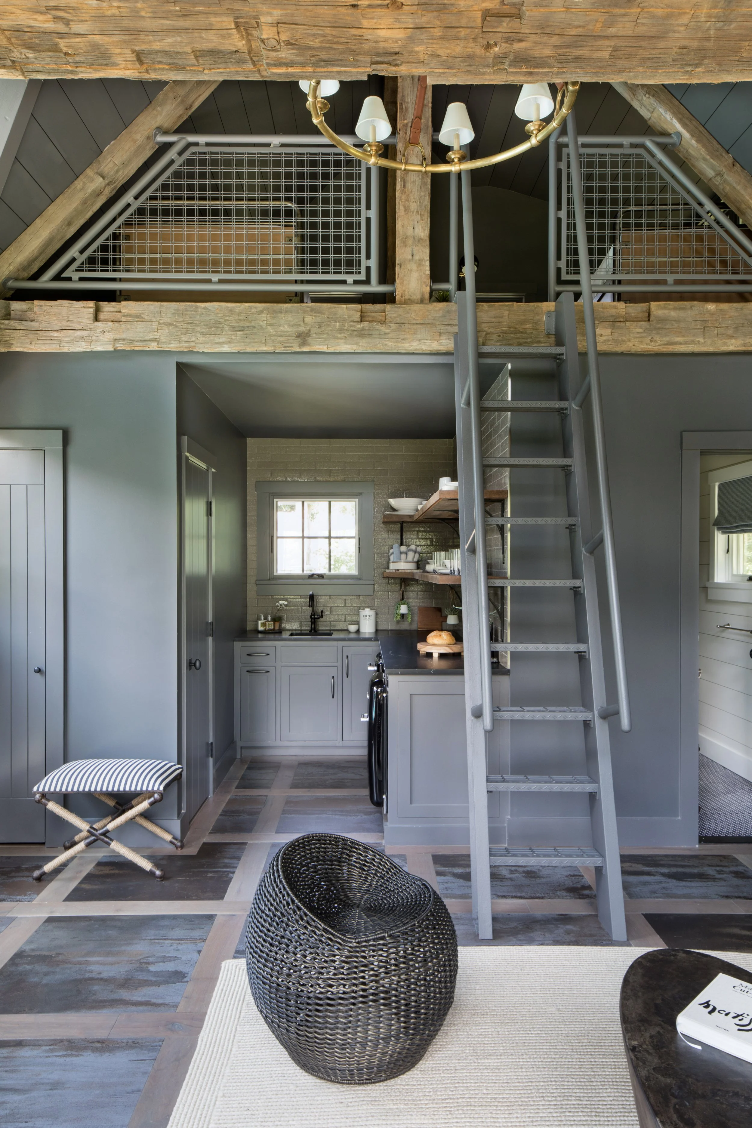 Interior view of a modern loft with a small kitchen, a staircase leading to a lofted upper area, and a seating area with a black woven chair and a textured rug. The loft railing has a metal grid design.