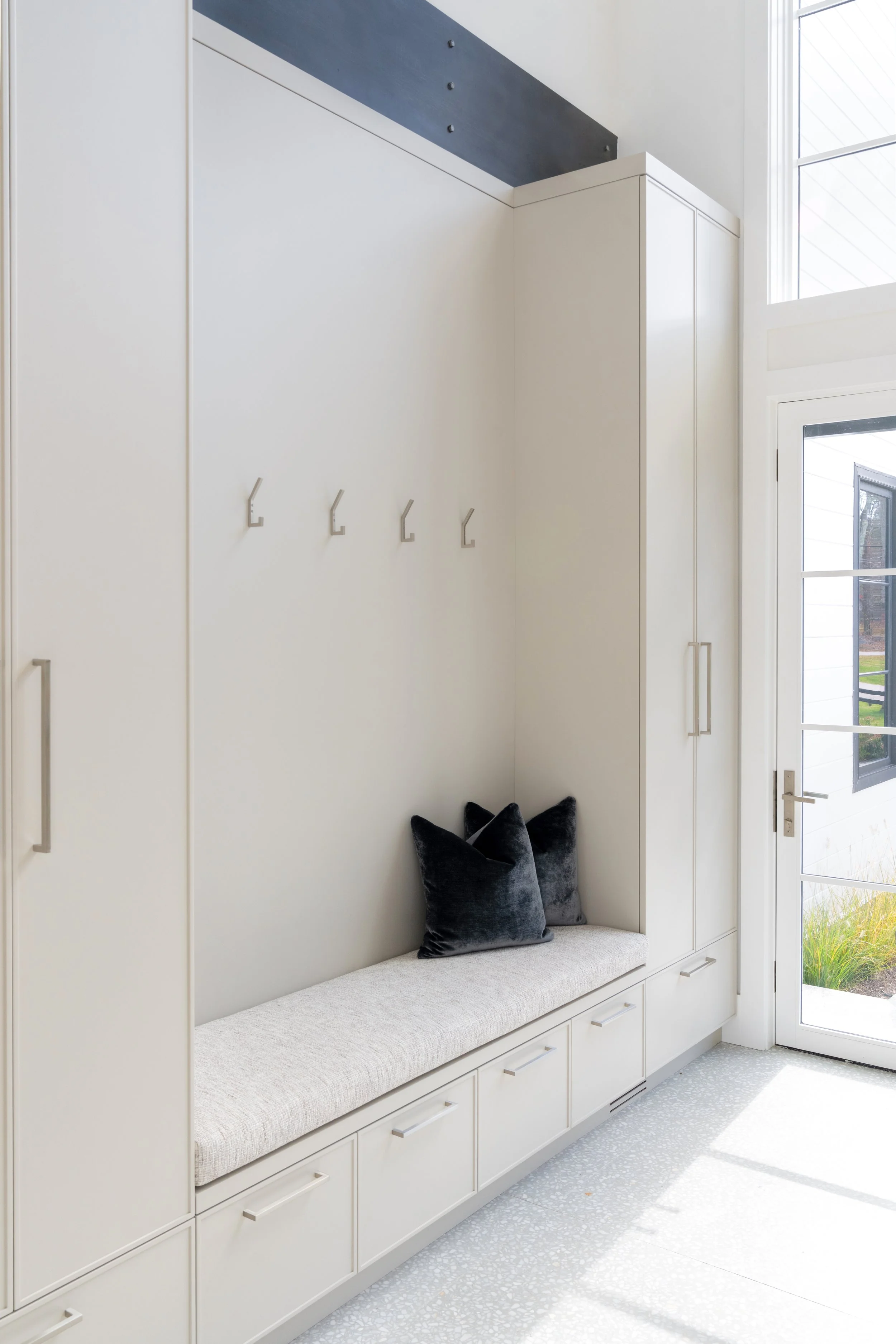 Entryway with white built-in storage, hooks on the wall, black pillows on a bench, and glass door leading outside.