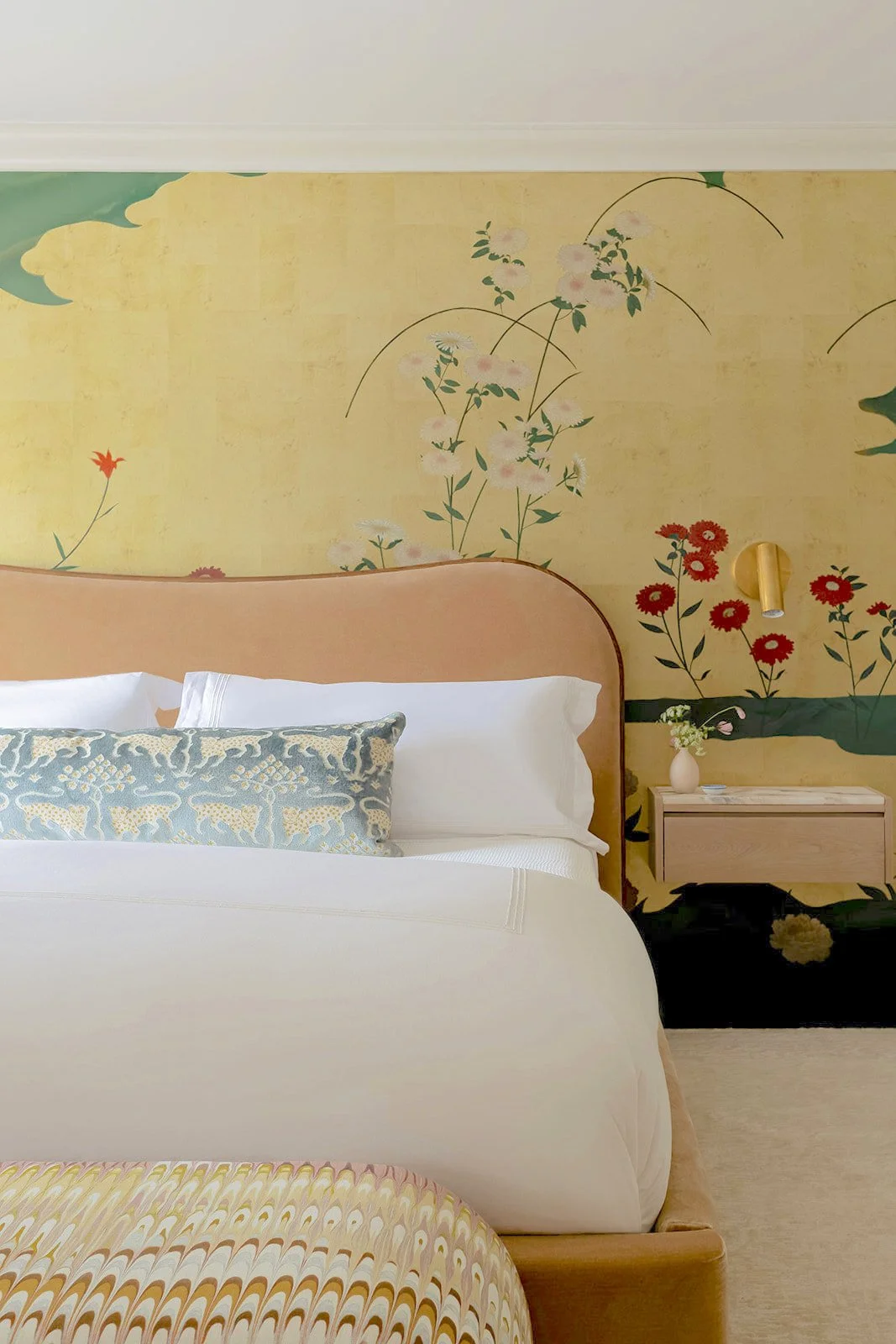 A bedroom with a large bed featuring white bedding and a decorative pillow with a leopard pattern, a light wood headboard, a beige nightstand with a small vase of flowers, a wall-mounted light fixture, and a floral mural with pastel-colored flowers o
