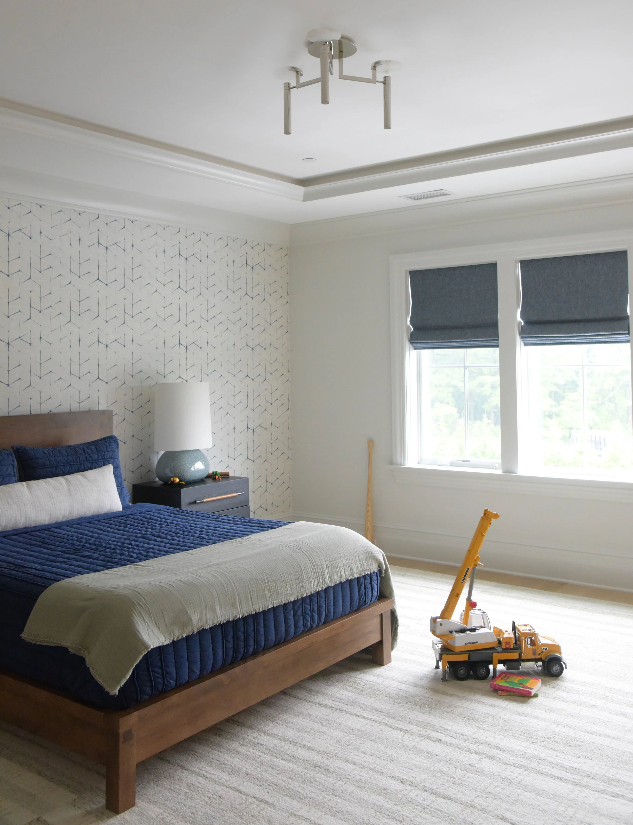 A bedroom with a wooden bed frame, blue bedding, cream throw blanket, white and blue pillows, a side table with a white lamp, window with blue shades, and a toy crane truck on the carpeted floor.
