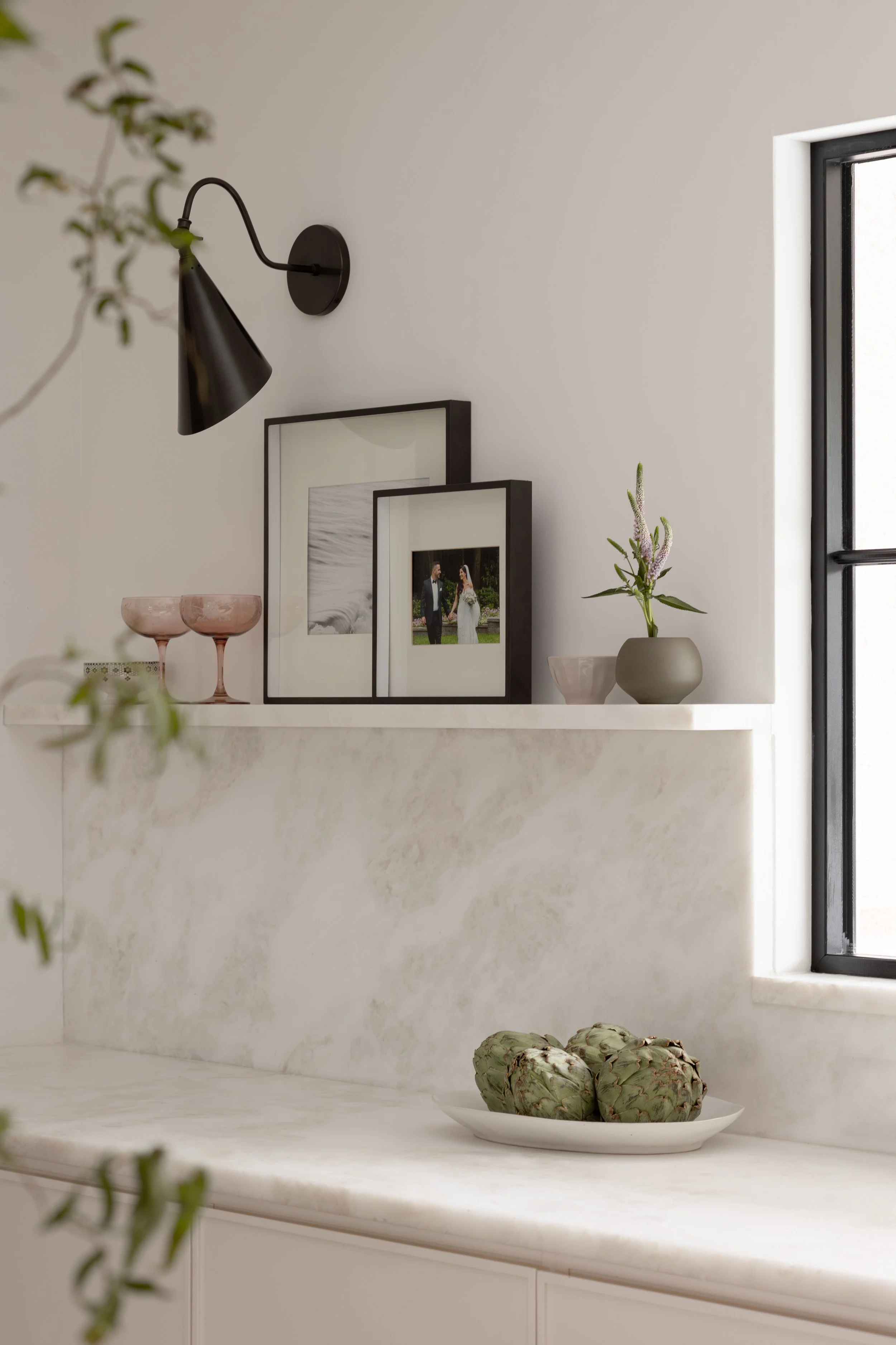 Decorative kitchen shelf with picture frames, vases, artichokes on a white marble countertop, and a window in the background.