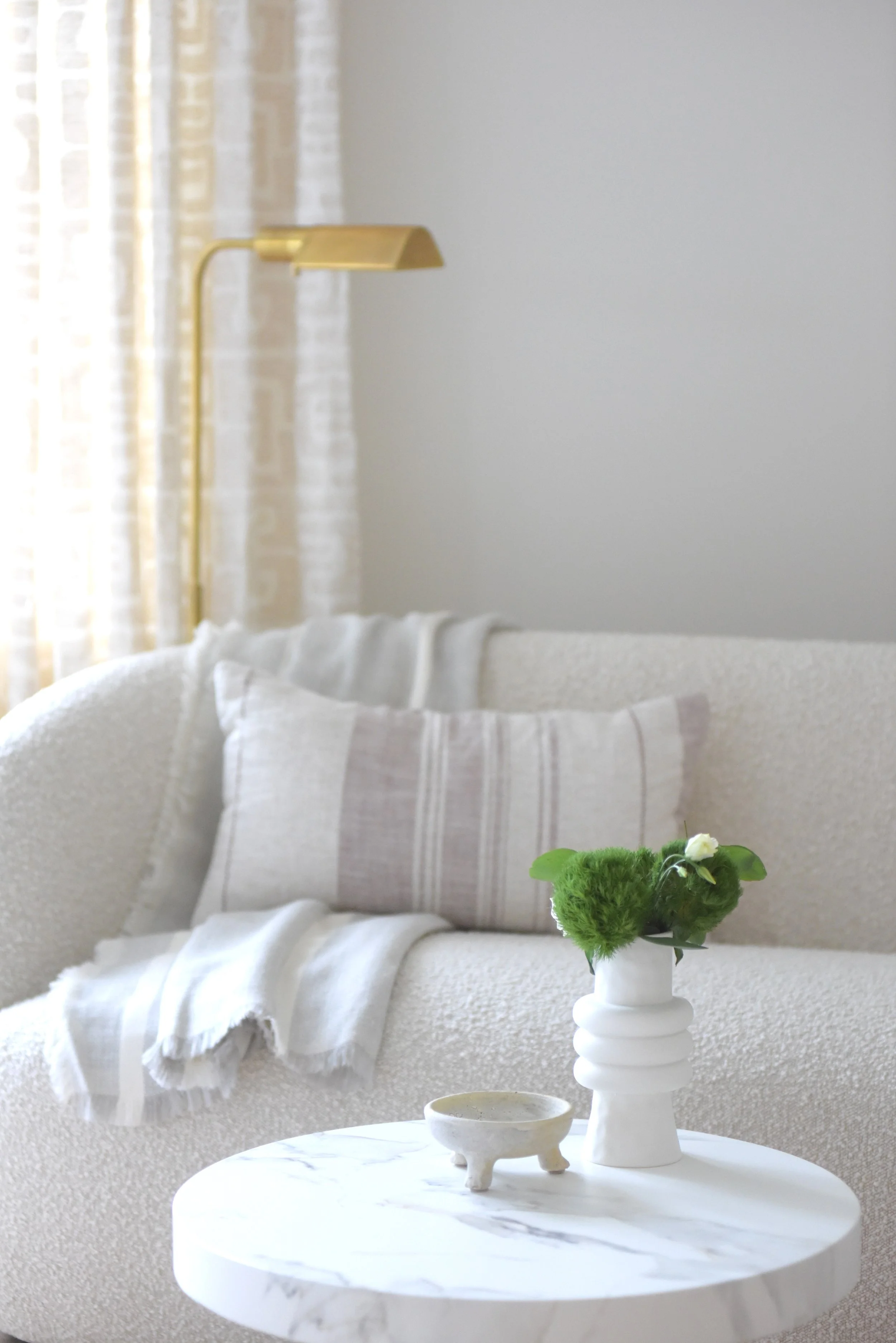 A cozy living room corner featuring a white textured sofa with light-colored pillows, a white throw blanket, a round marble side table with a white vase holding green flowers, and a small white decorative dish. In the background, a gold wall-mounted 