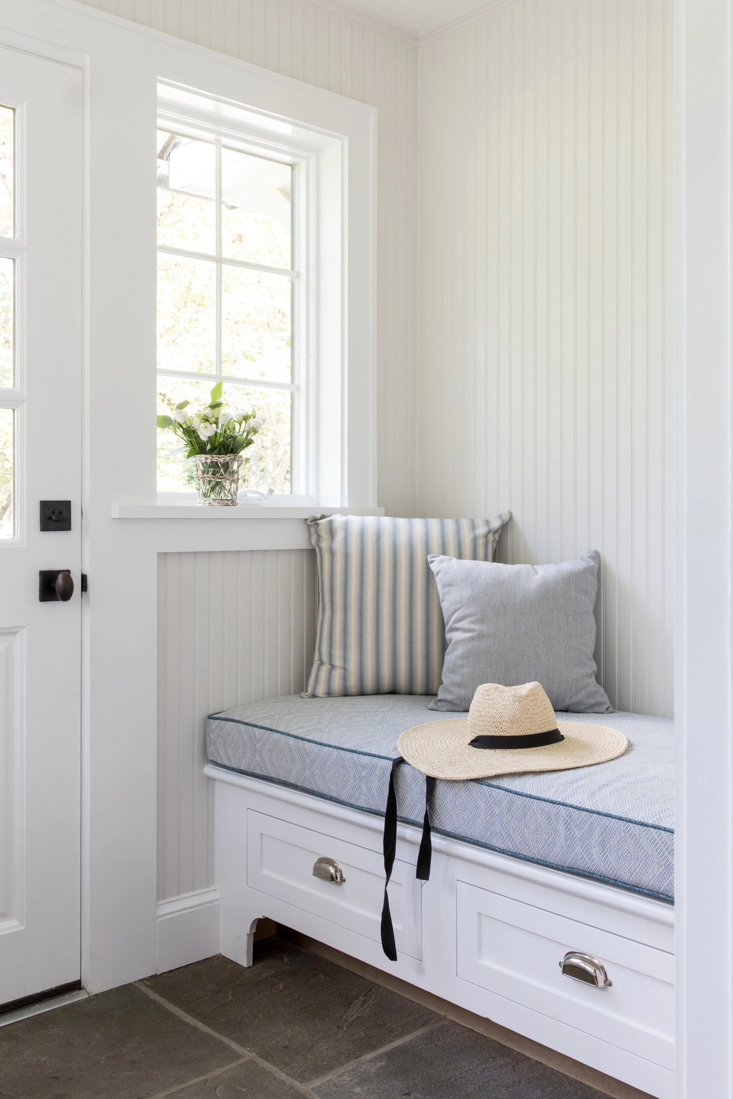 A cozy white window seat with two pillows, a straw hat with a black ribbon, and a vase of white flowers on the window ledge, in a bright room with white paneled walls.
