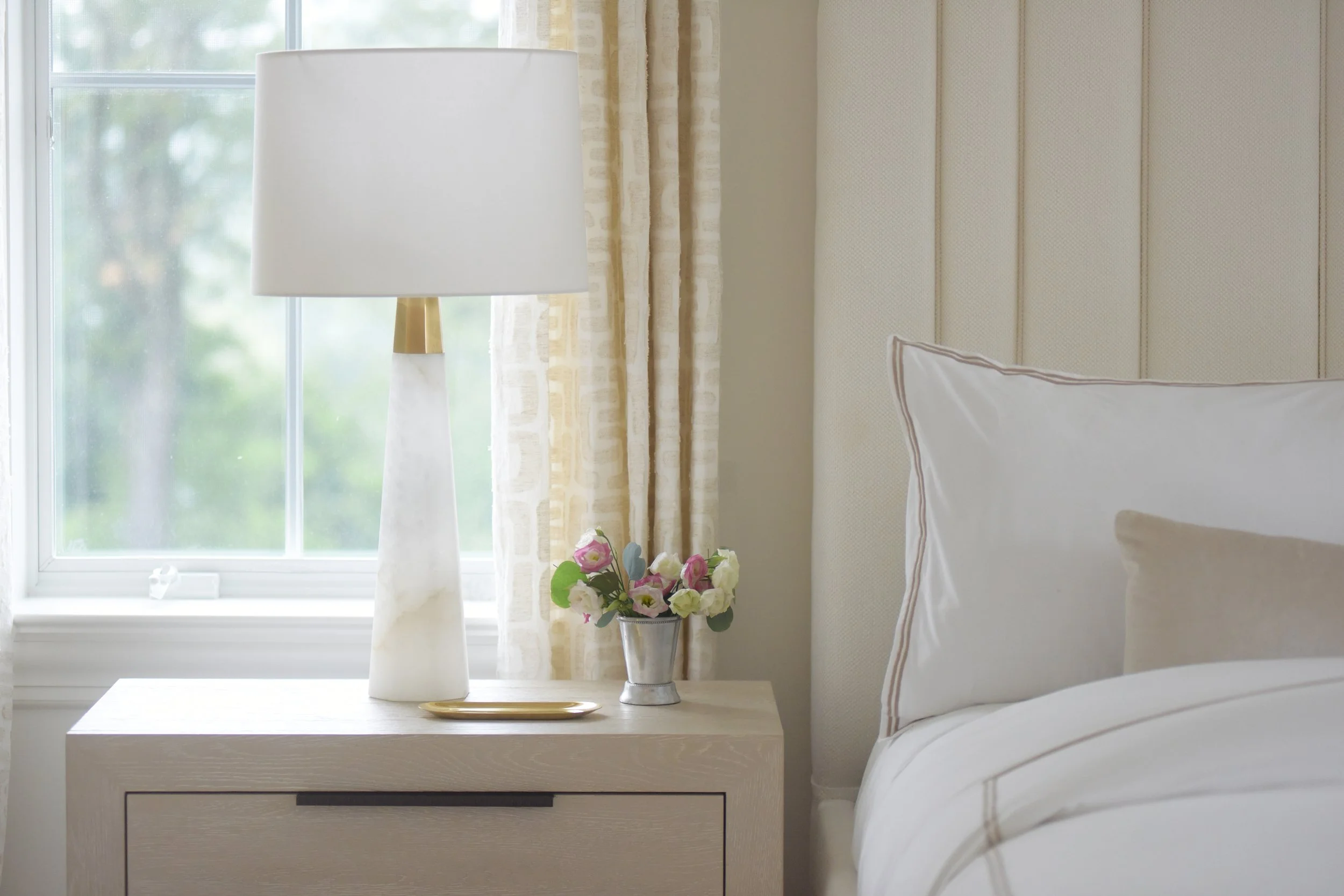 Bedroom with a bedside table, a lamp, a window, curtains, and a bed with pillows and a headboard.