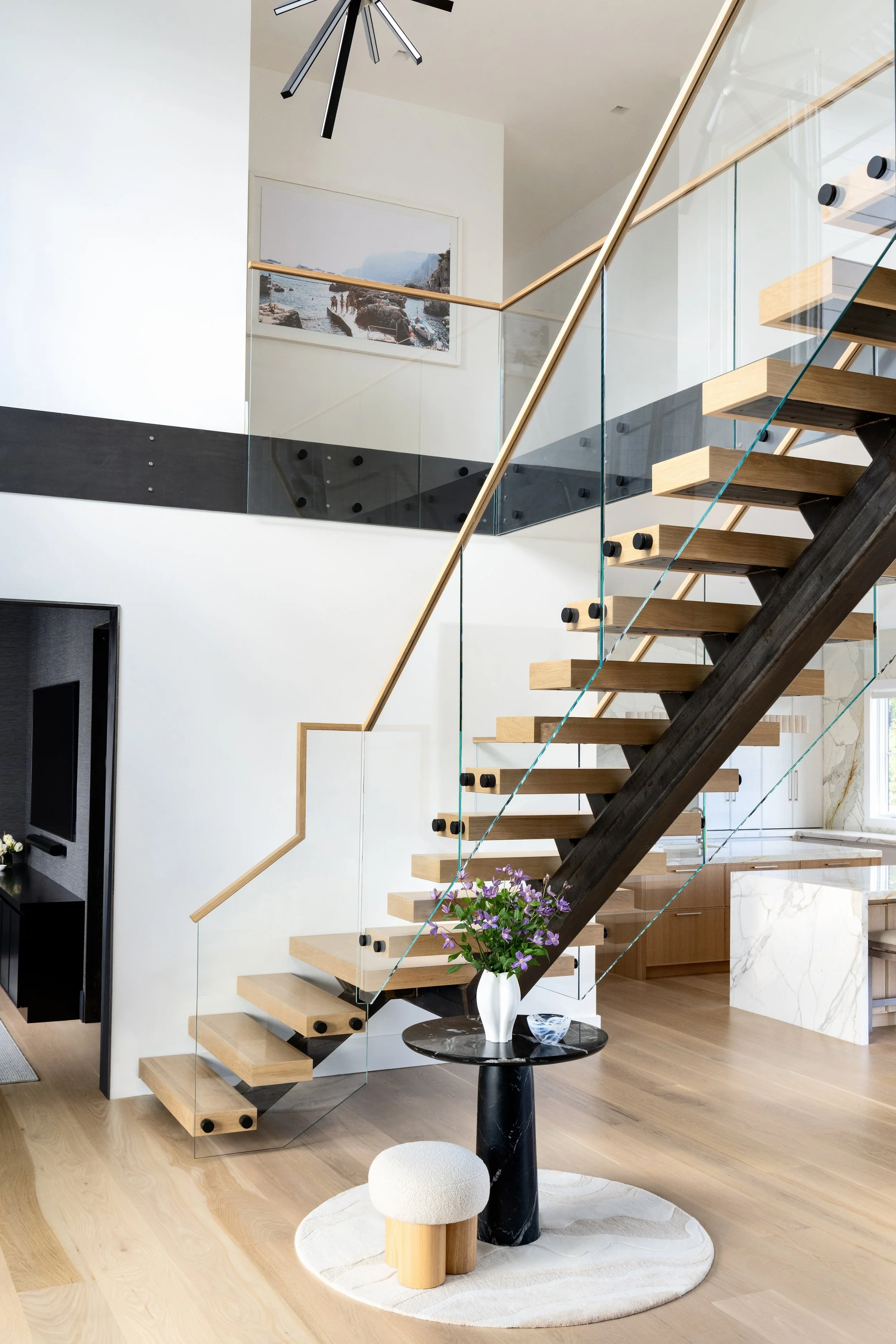 Modern interior with a staircase, black and wooden steps, glass railing, and a small round table with a white vase of purple flowers.