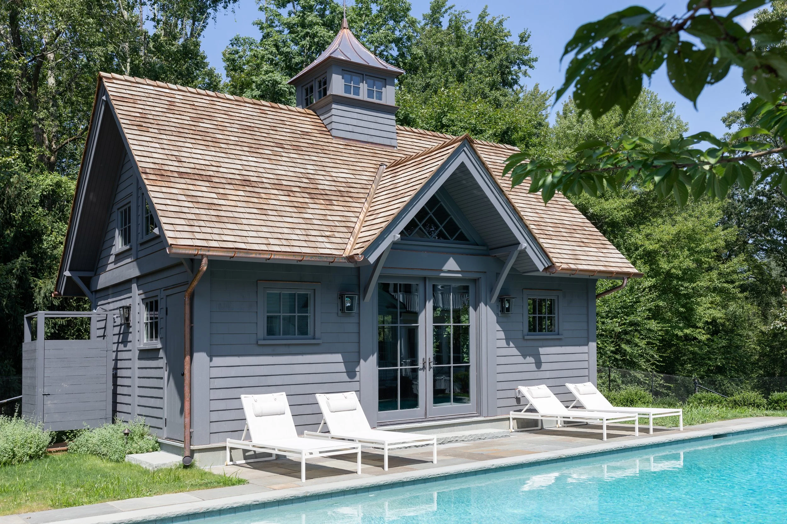 A cozy blue house with a wooden shingle roof, large glass doors, and small windows, situated next to a swimming pool with white lounge chairs under a sunny sky surrounded by green trees and foliage.