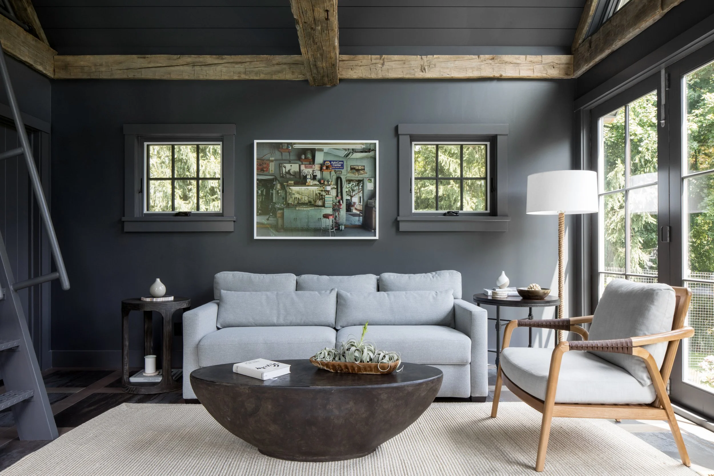 Living room with gray sofa, wooden armchair, large window, dark walls, ceiling beams, and artwork on the wall.