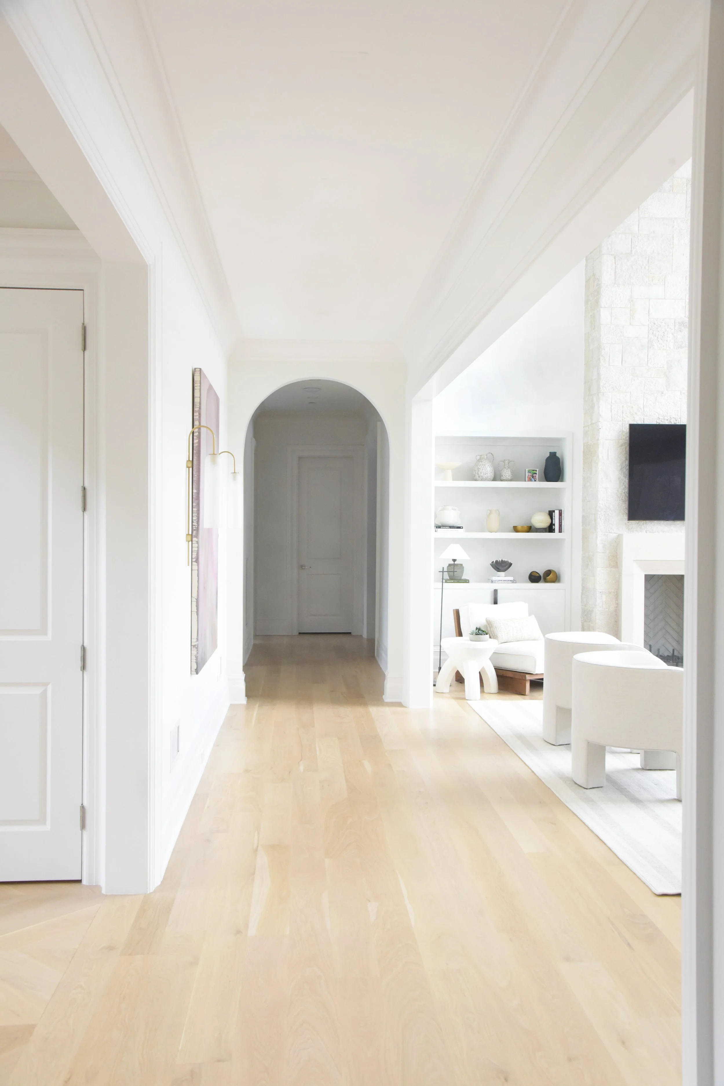 Bright, white interior living space with light wood floors, built-in shelves with decorative items, white sitting area, and a TV mounted on a stone fireplace wall.