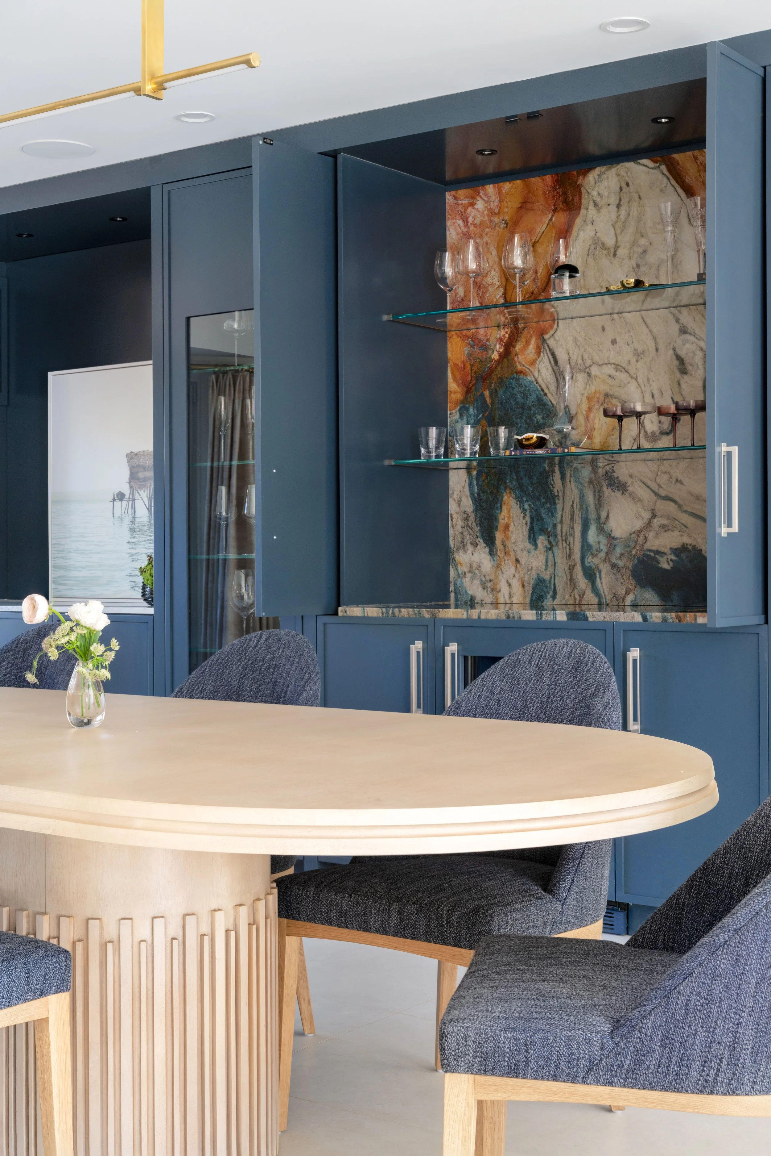 Modern dining room with a round light wood table, dark gray upholstered chairs, and a built-in blue cabinet featuring a marble backsplash with glass shelves and glassware.