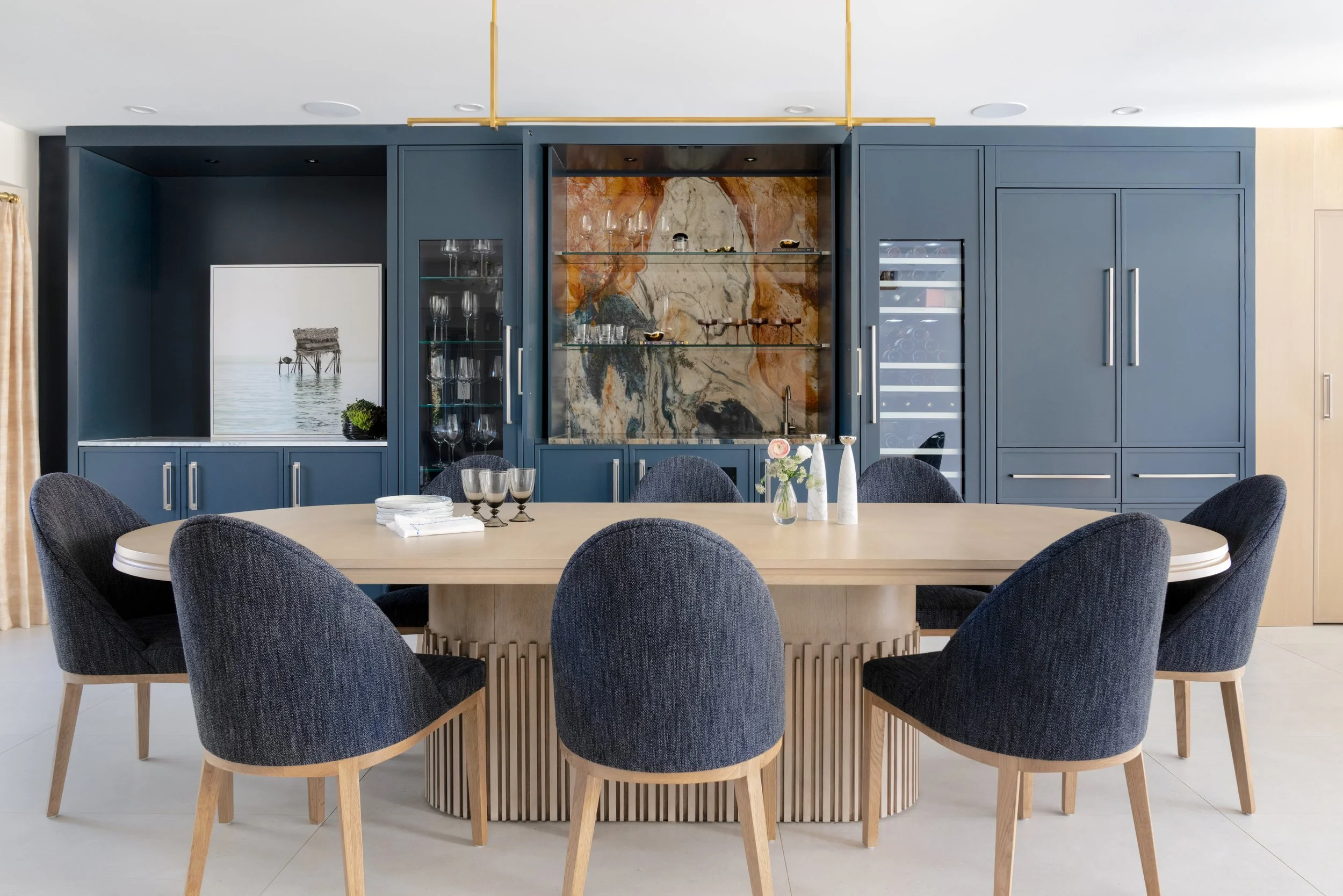 Modern dining room with a light wood oval table, eight dark blue upholstered chairs, and a built-in blue cabinet with glass shelves and a colorful stone backsplash.
