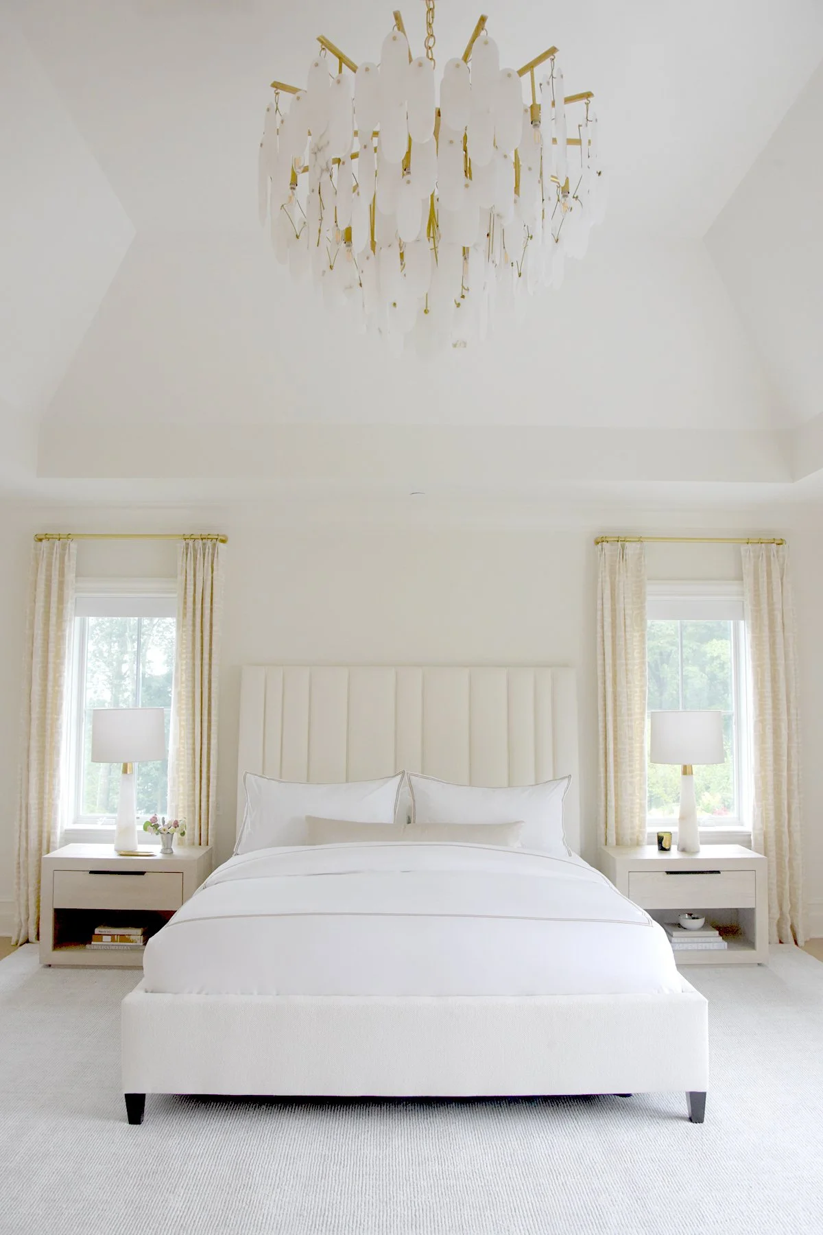 A bright, minimalistic bedroom with a white bed, two nightstands with lamps, and windows with cream curtains. A chandelier hangs from the ceiling.