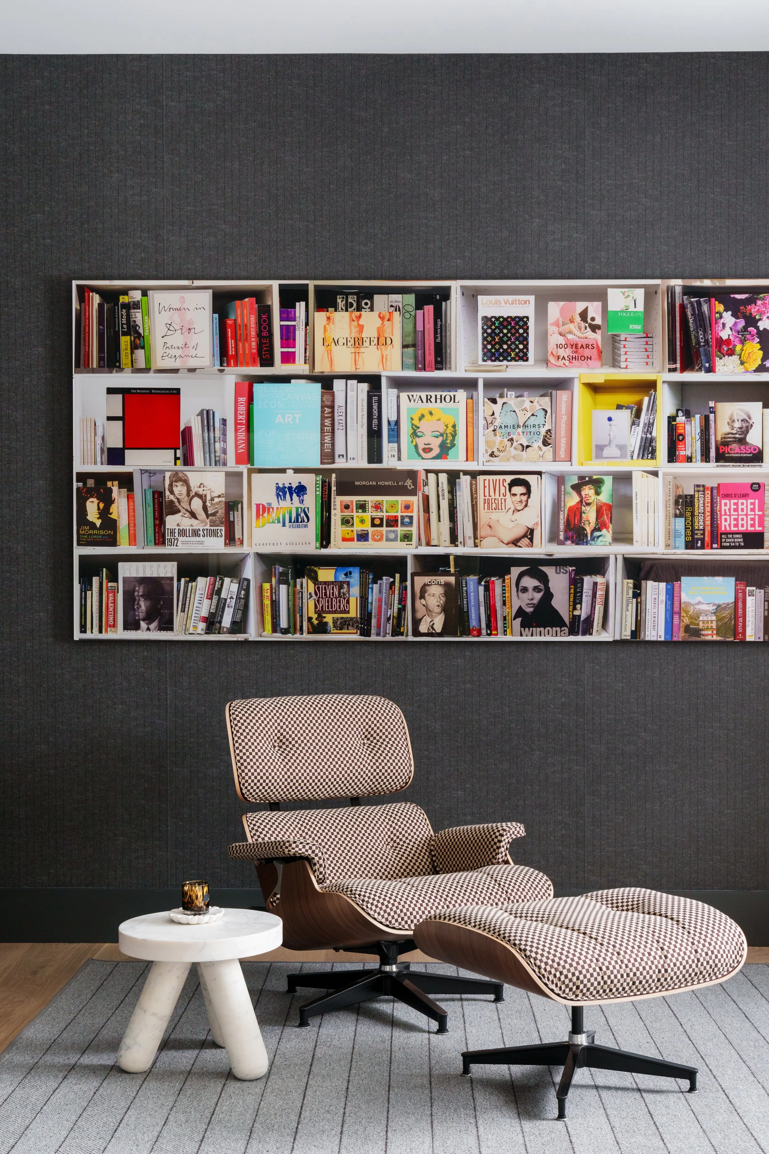 A modern reading nook with a black wall, a white bookshelf filled with books, a houndstooth-patterned lounge chair with matching ottoman, a small round white marble side table, and a glass of whiskey on the table.