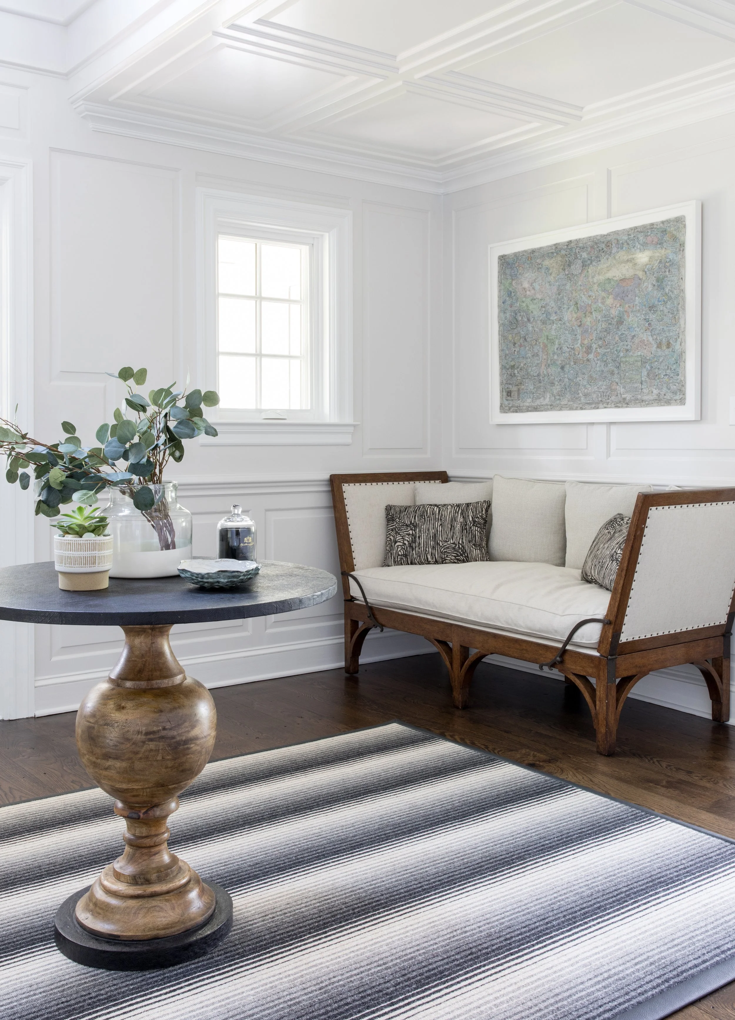 A cozy living room corner with a white paneled wall, a small window, a framed artwork, a wooden bench with cushions, a round wooden table with decorative items, and a striped rug on dark hardwood floor.