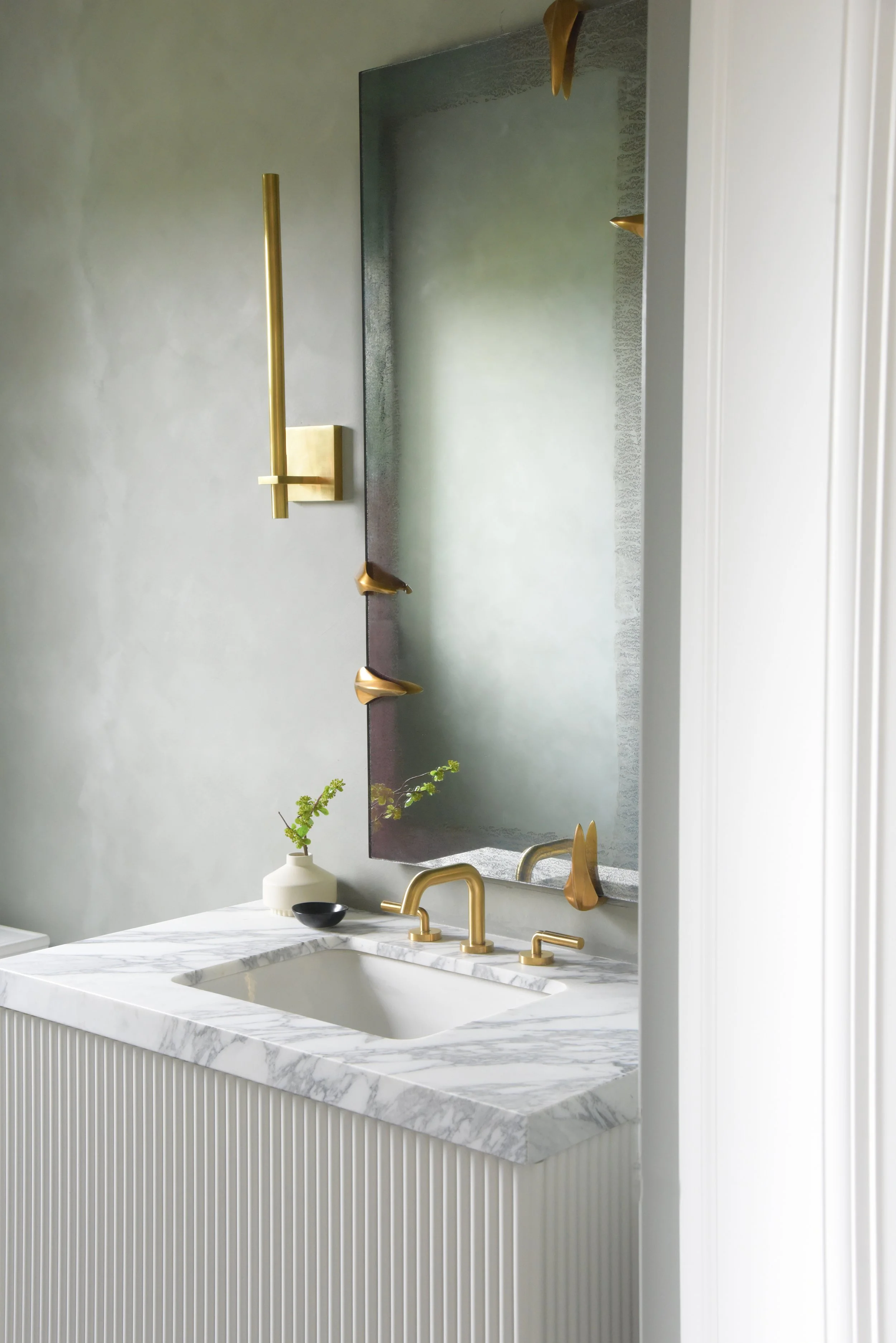 Bathroom vanity with a marble countertop, gold fixtures, and a large mirror with decorative gold accents. A small vase with greenery and a black bowl are on the countertop.