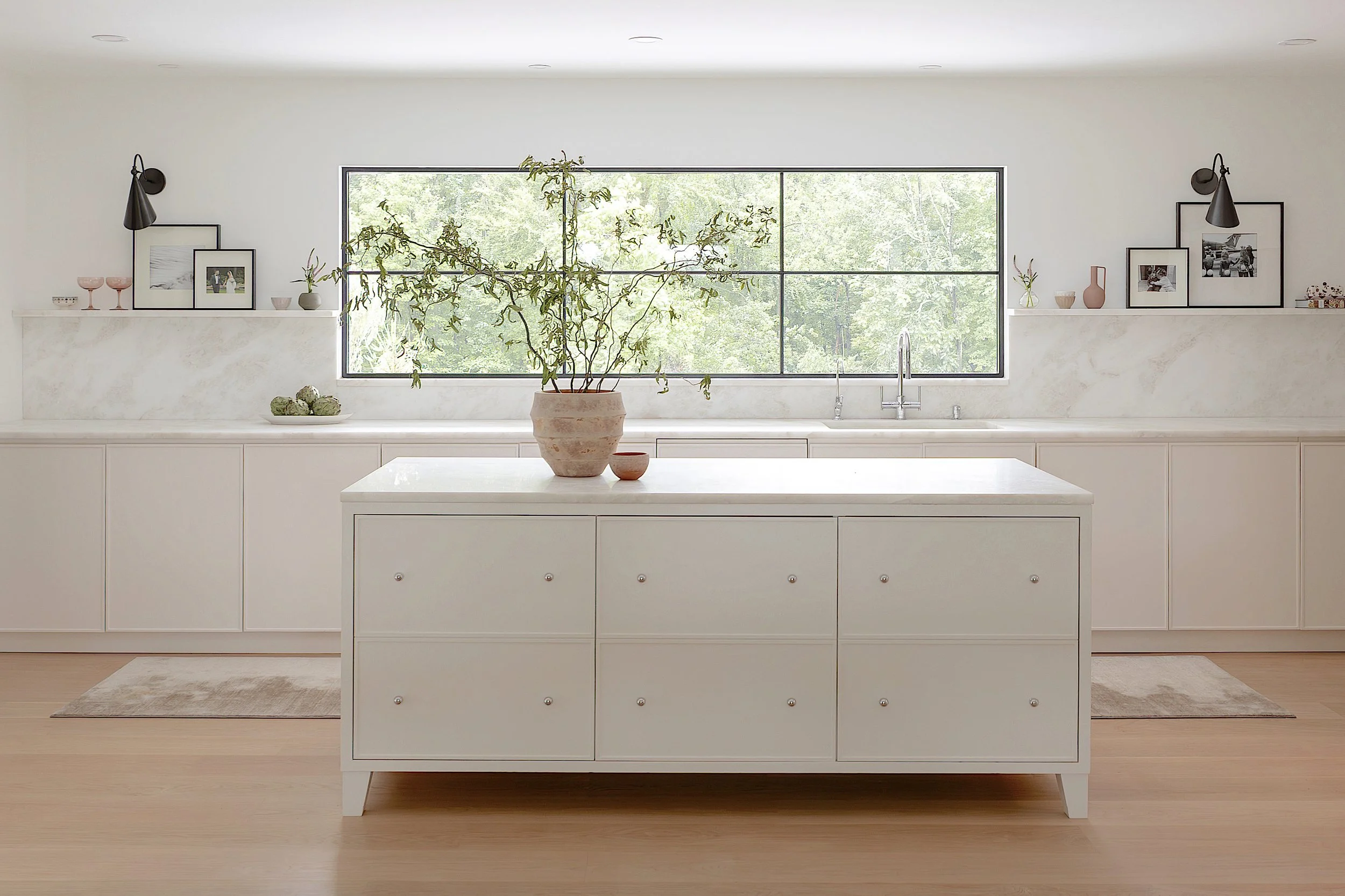Bright, minimalist kitchen with white cabinets, a large window showing greenery outside, a white counter and island with a potted plant, and decorative items on shelves.