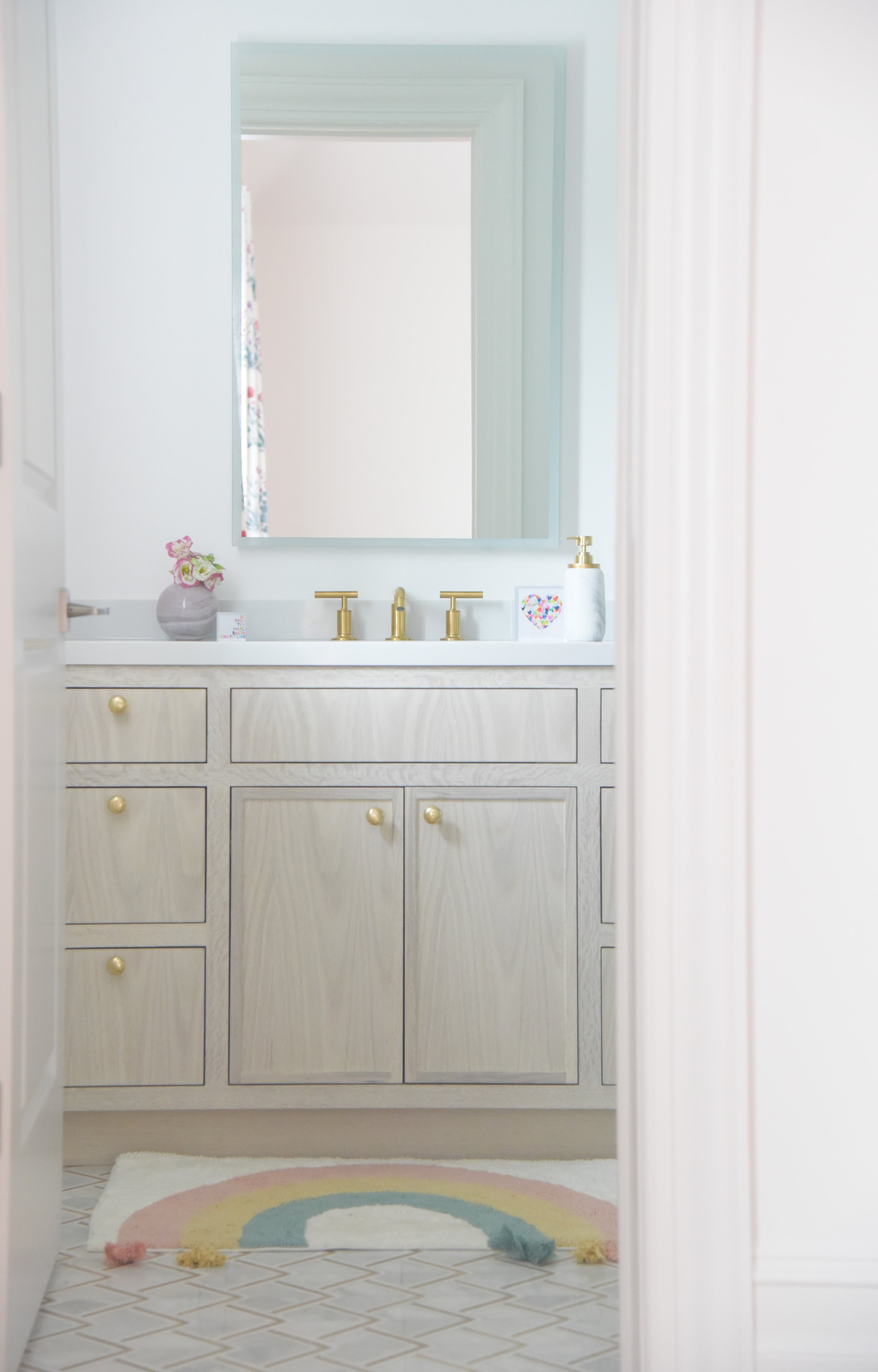 A bathroom sink area with a large mirror, gold faucet fixtures, light-colored wooden cabinets, a vase with pink flowers, a soap dispenser, and a small heart-shaped decoration. A multicolored rainbow-shaped rug is on the floor.