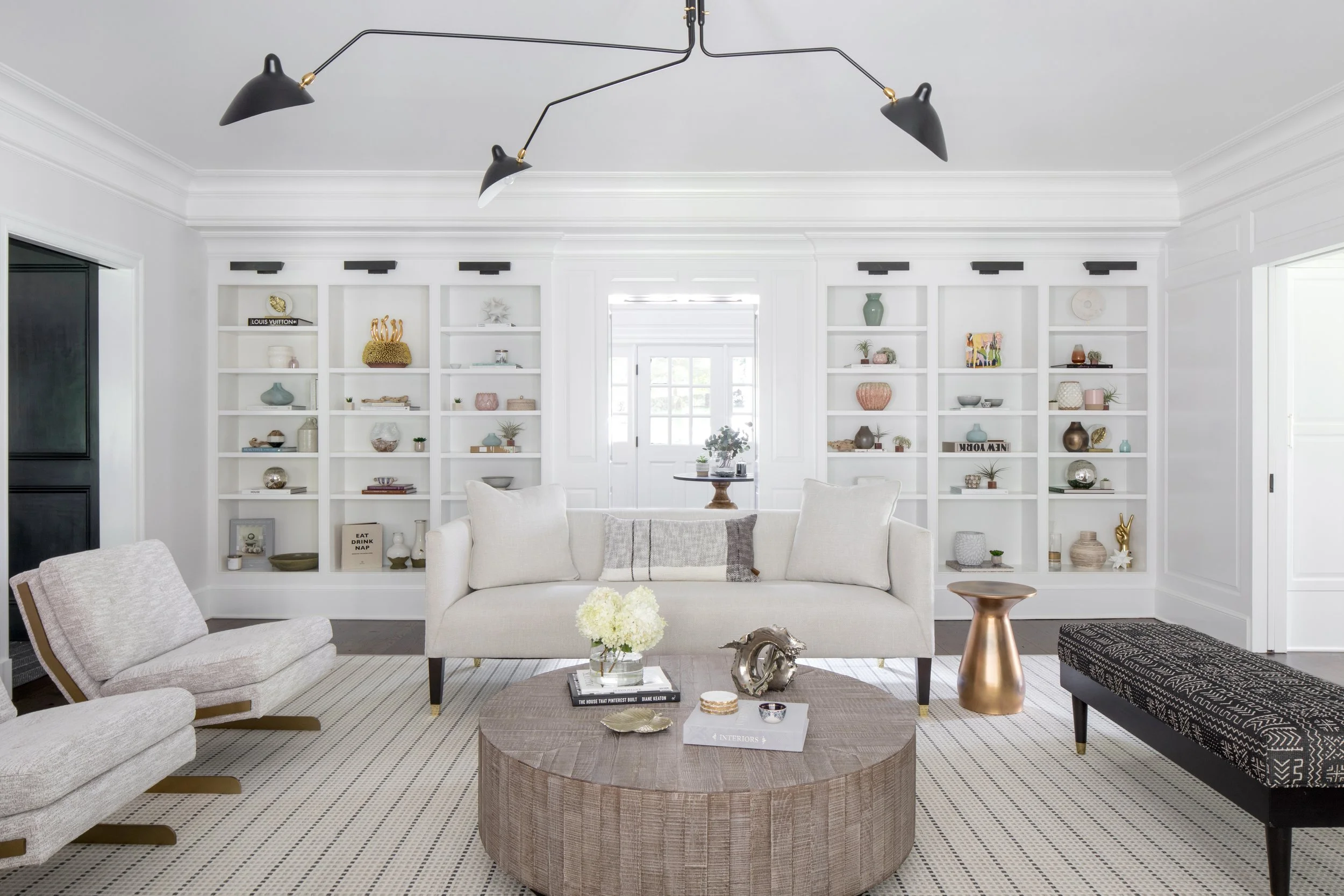 Living room with white sofa, beige armchair, black and white patterned bench, round wood coffee table, white built-in bookshelves, and modern black ceiling light fixture.