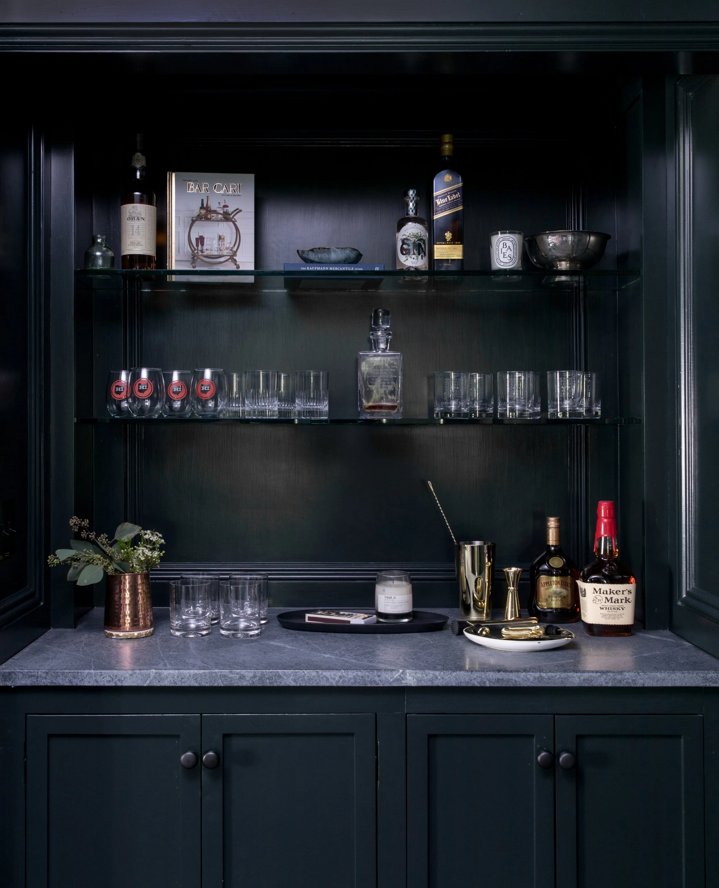 A dark green bar cabinet with a gray countertop, barware, bottles, glasses, a candle, and a small bouquet of flowers in a copper vase.