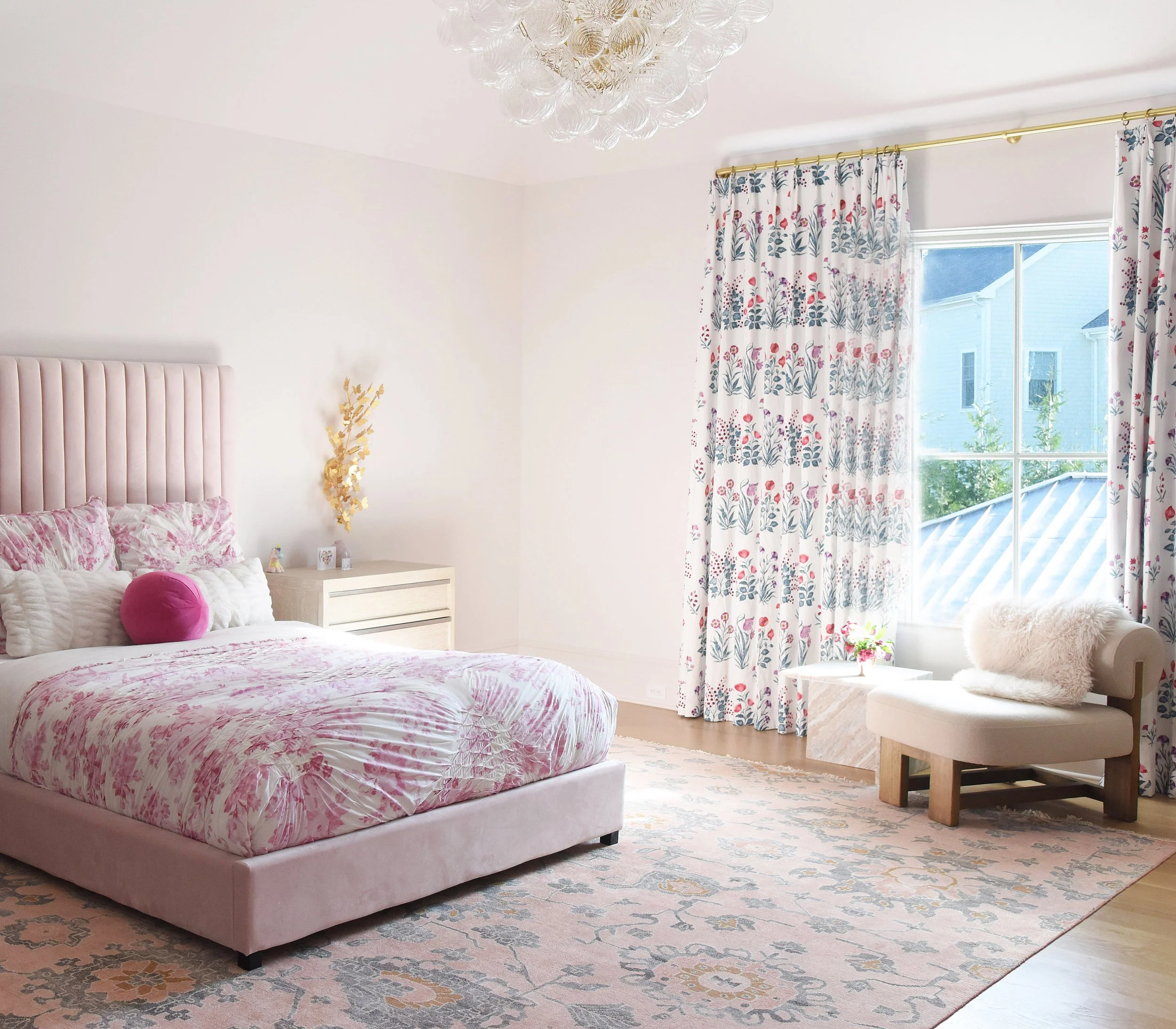 Bright bedroom with pink and white bedding, pink velvet headboard, floral curtains, white chair with fur pillow, cream nightstand, decorative wall piece, and a large window letting in natural light.