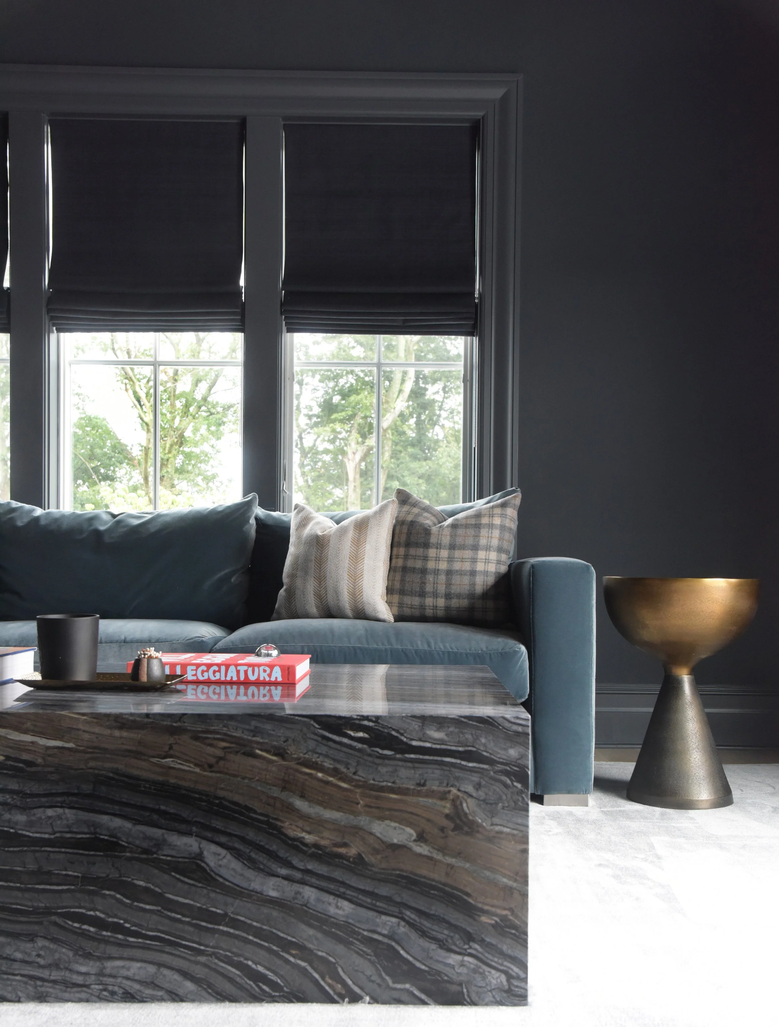 Living room featuring a blue sofa with plaid and cream pillows, a marble coffee table, black window blinds, a gold side table, and a large window with trees visible outside.