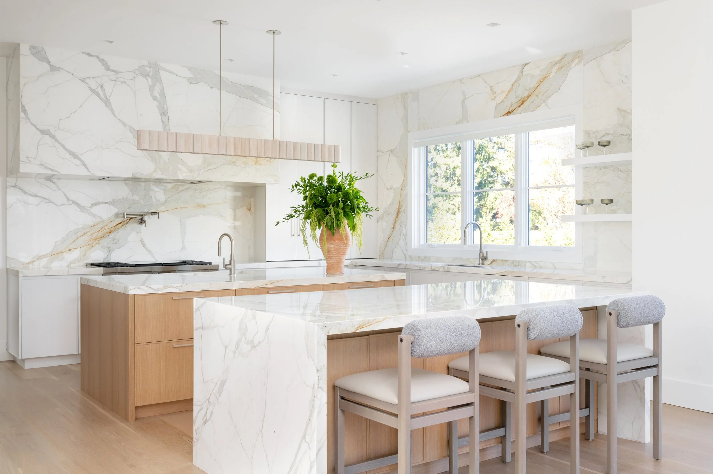 Modern kitchen with marble countertops, a large island, white cabinetry, a large window, a potted plant on the island, and light wood bar stools.