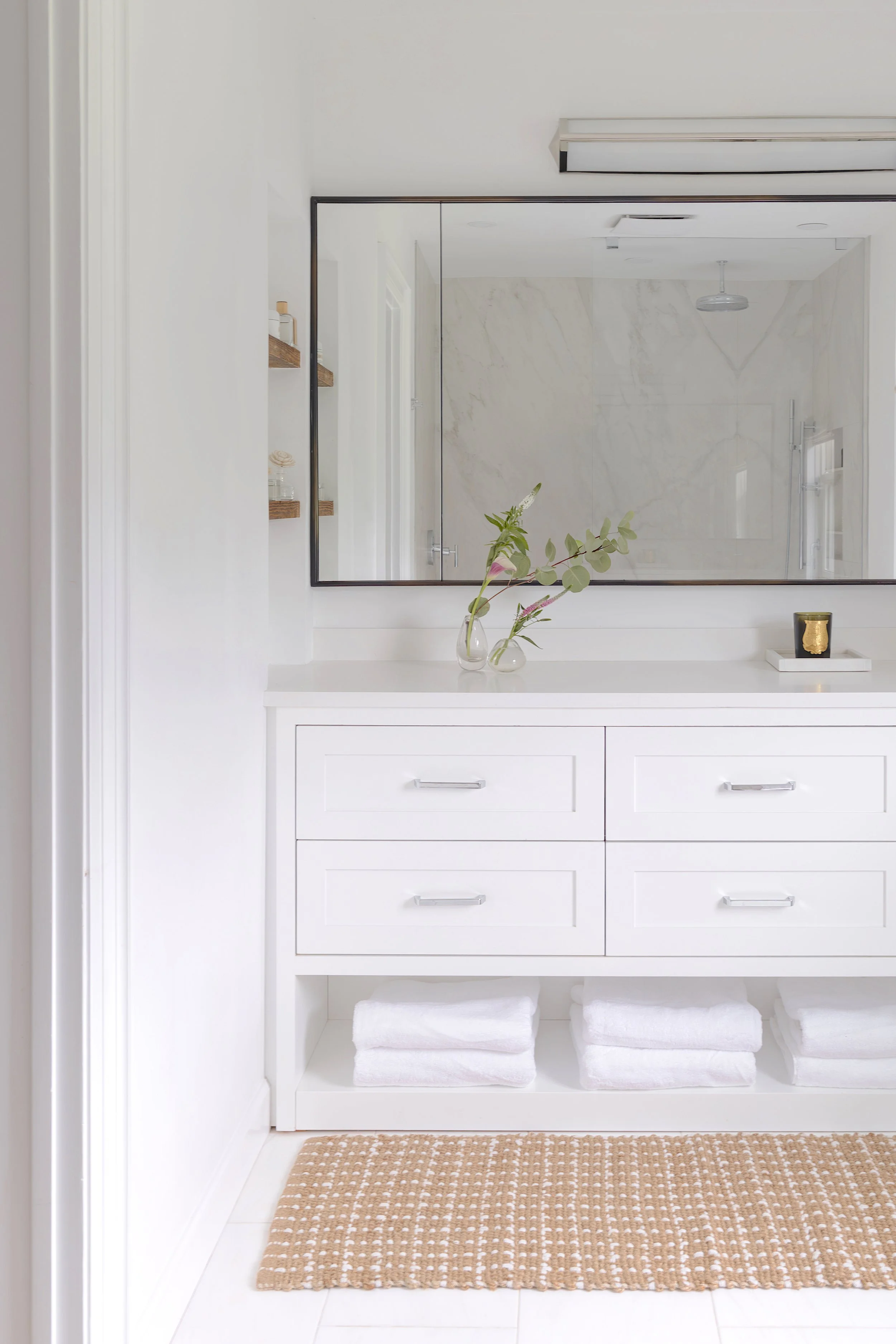 White bathroom vanity with drawers, mirror, and shelves, decorated with a vase of green and pink flowers, beige woven rug, and towels underneath.