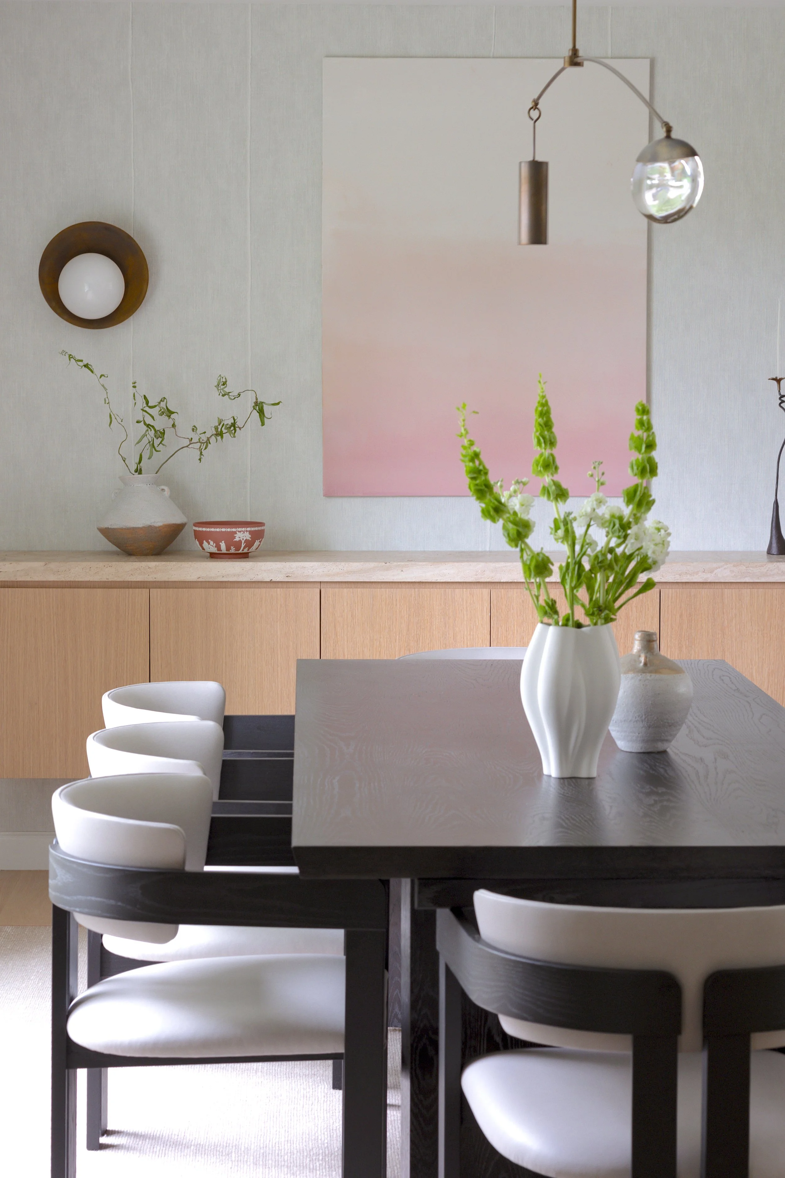 A dining room with a dark wood table, white chairs, and decorative vases filled with green and white flowers, with a wood sideboard, wall art, and contemporary lighting fixtures in the background.