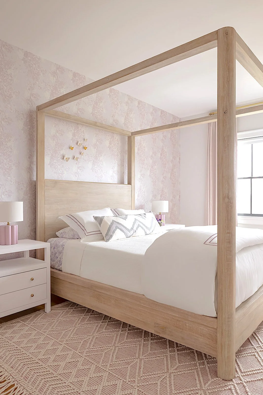 A bedroom with a light wood four-poster bed, white bedding, matching pillows, and pink lamps on white nightstands. The room features a pink patterned rug, floral wallpaper, and a window with pink curtains.