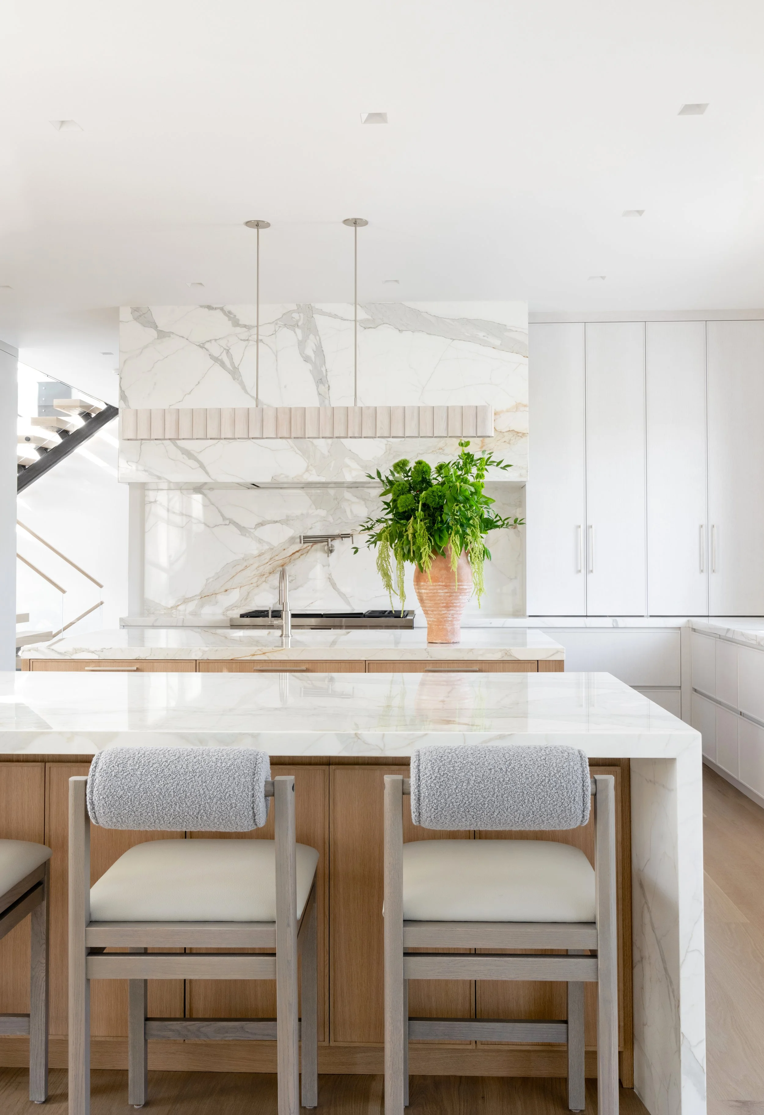 Modern kitchen with white marble countertops, a large potted green plant, and beige bar stools with light wood frames.